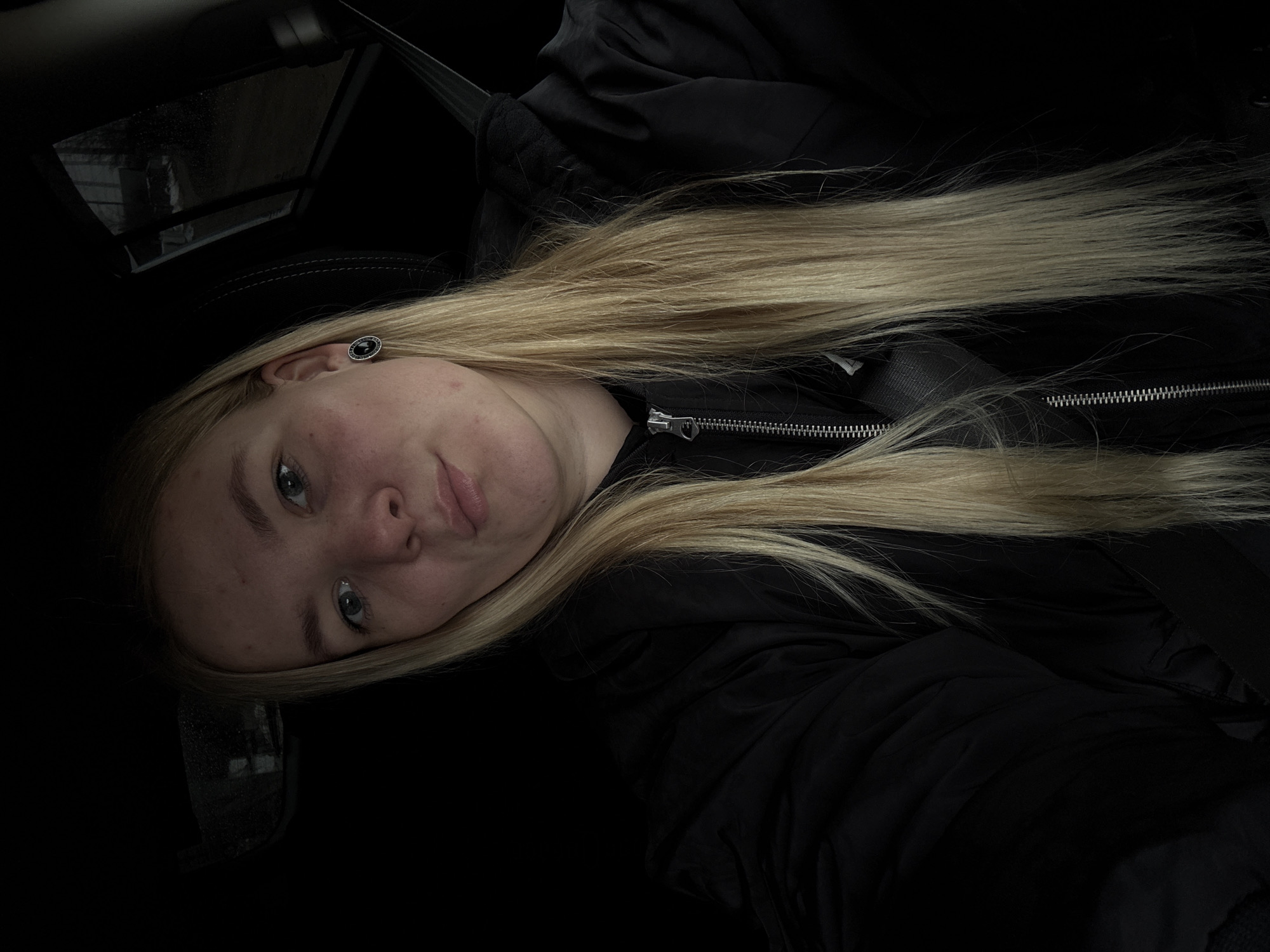 A young person with blonde hair, light complexion, and blue eyes is reclining, possibly in a car, as evidenced by the seatbelt visible in the upper portion of the frame. The individual is wearing a black jacket and has an earring in their left ear. The photograph appears to have been taken indoors, possibly within a vehicle, and shows a portion of a window on the left side. The location of Chaikovsky, Russia is not visually apparent.