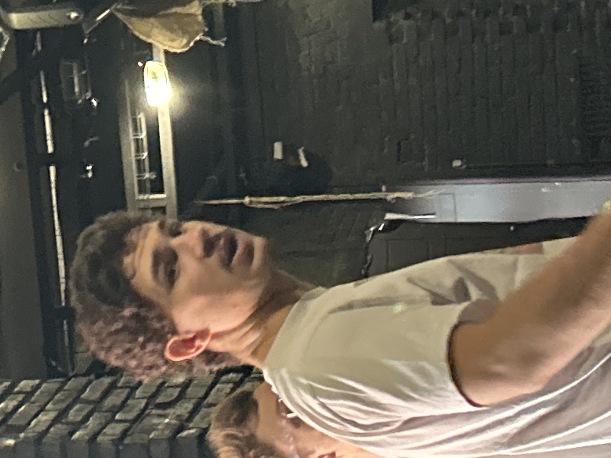 A young man with curly brown hair and fair skin is the primary subject of this image, which appears to be taken at night. He is wearing a light-colored t-shirt. His head is tilted back and his mouth is open, suggesting he might be speaking, laughing, or singing. His expression is animated and he has a light-colored ear visible. He is positioned in the foreground of the image, with a blurred background that includes what looks like a tiled surface below him and a dark, textured wall behind. There are also hints of industrial elements in the background, like metal structures and a light bulb. The lighting is dim, indicating it is likely nighttime.
