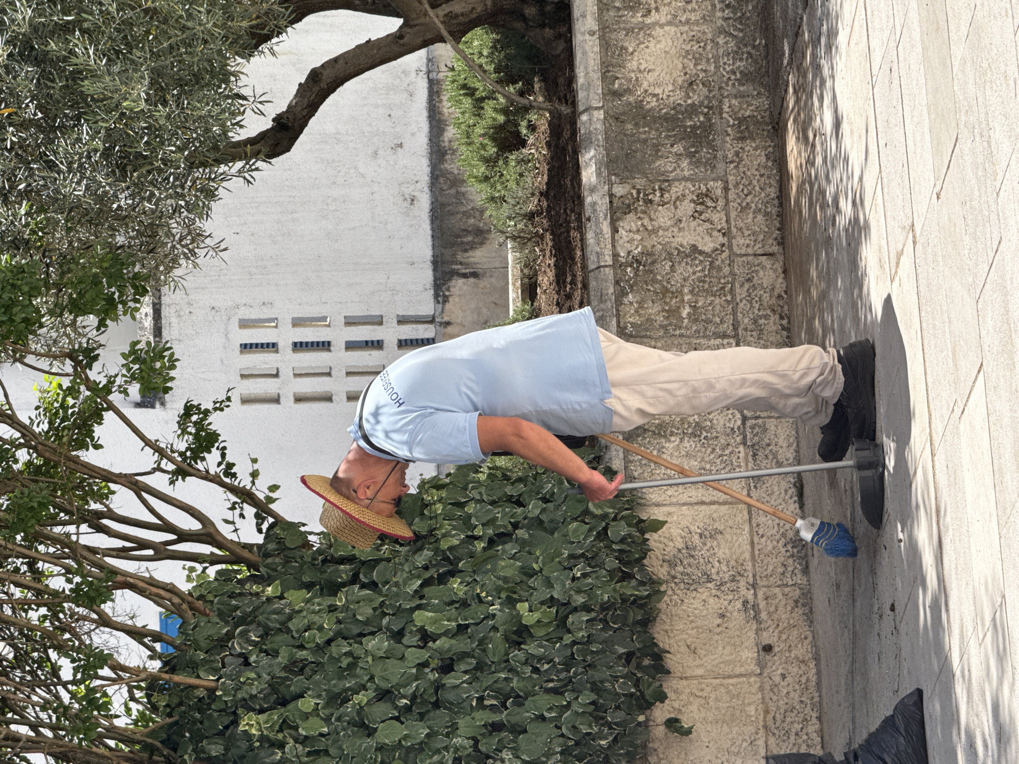 A man in a straw hat, light blue shirt, and khaki pants sweeps a stone paved area in Budva, Montenegro. He holds a blue-bristled broom. A wall and greenery are adjacent to the paved area. The white wall has small rectangular openings. Trees are present.