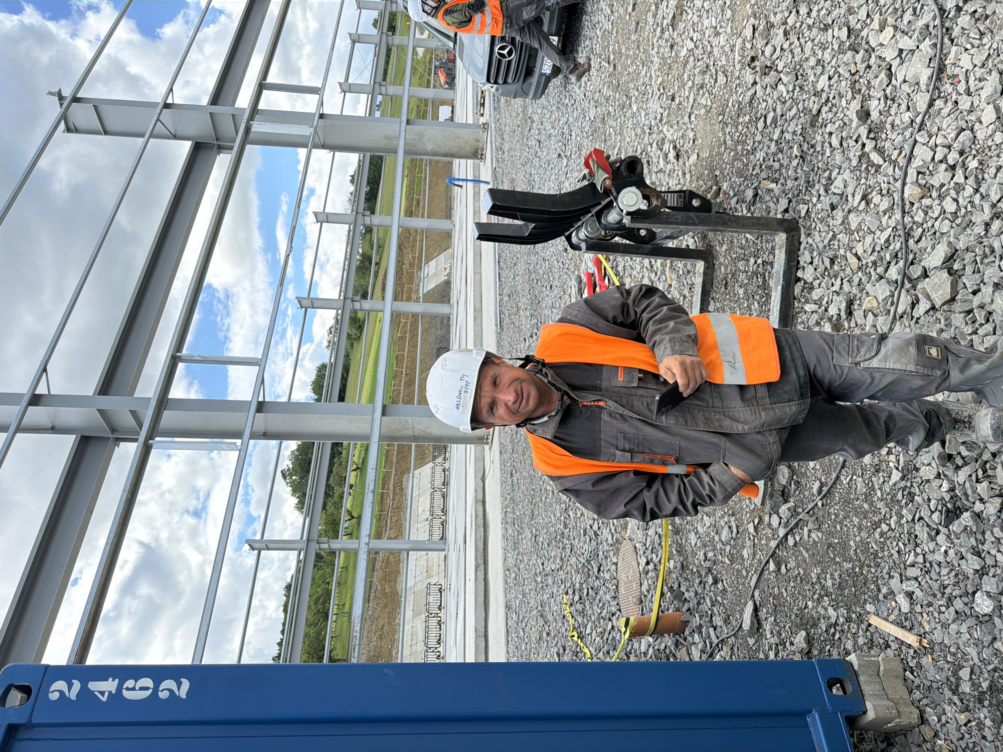 A man wearing a hard hat and an orange safety vest stands in front of a large blue shipping container at a construction site in Stróża, Poland. He holds a smartphone in his left hand and his right hand is in his pocket. The construction site features a steel structure with large beams and a gravel-covered ground. In the background, a van and some construction equipment are visible. The sky is partly cloudy.