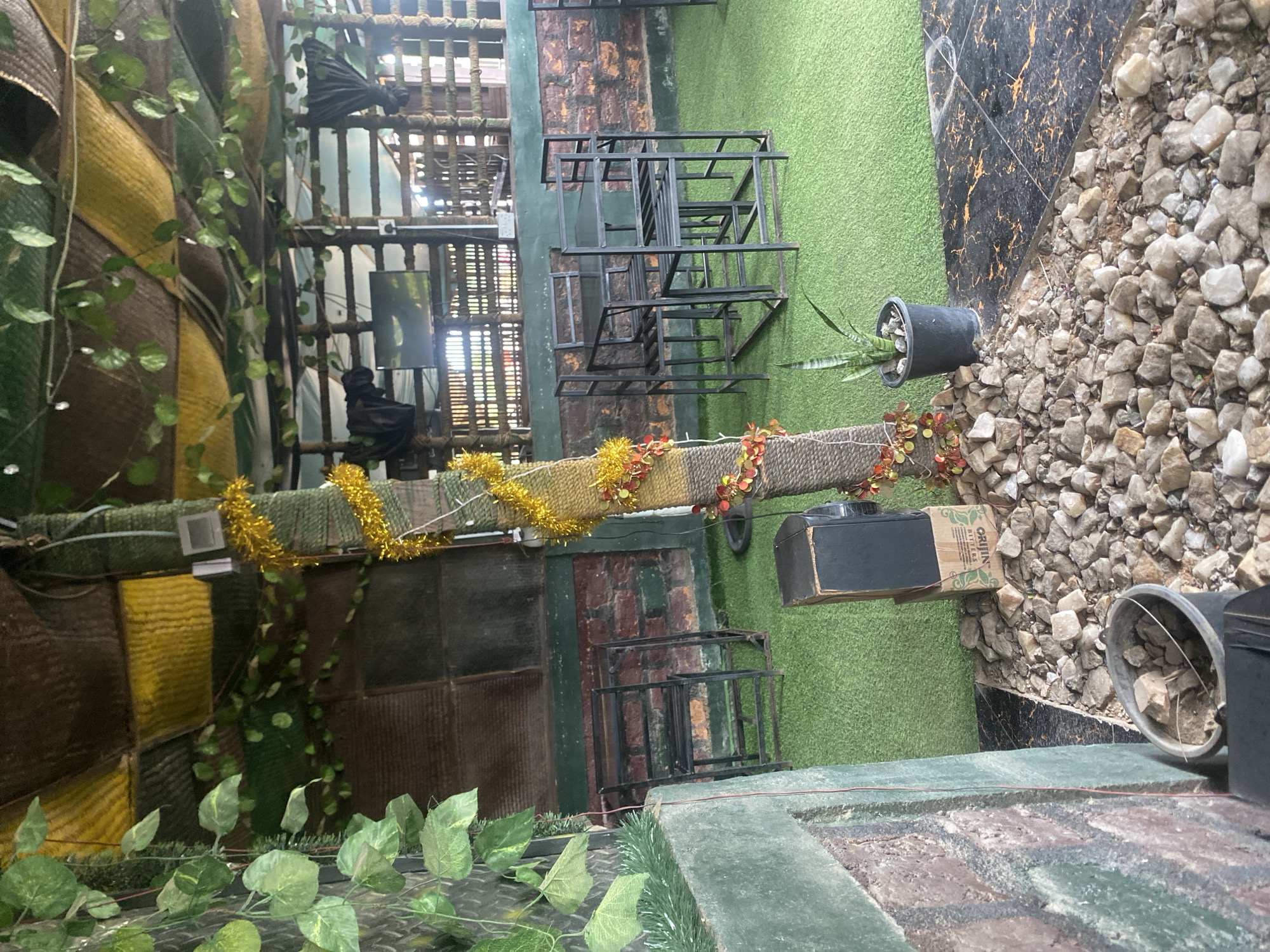 The image depicts an outdoor seating area at a restaurant or bar in Jalingo, Nigeria. The space is characterized by a rustic, natural aesthetic. Artificial turf covers the ground, and an overhead structure is draped with yellow and brown fabric, creating a canopy. Artificial green vines hang from the canopy and from a central, tree-like pillar adorned with gold tinsel and red, leaf-like decorations.
Several black metal tables and chairs are arranged on the turf, suggesting seating for patrons. A black speaker sits on a cardboard box near the base of the decorated pillar. A potted snake plant is also visible. The background reveals a structure with wooden slatted walls and a brick wall, hinting at the building's construction. The overall impression is that of a casual, possibly themed, outdoor dining or relaxation space.