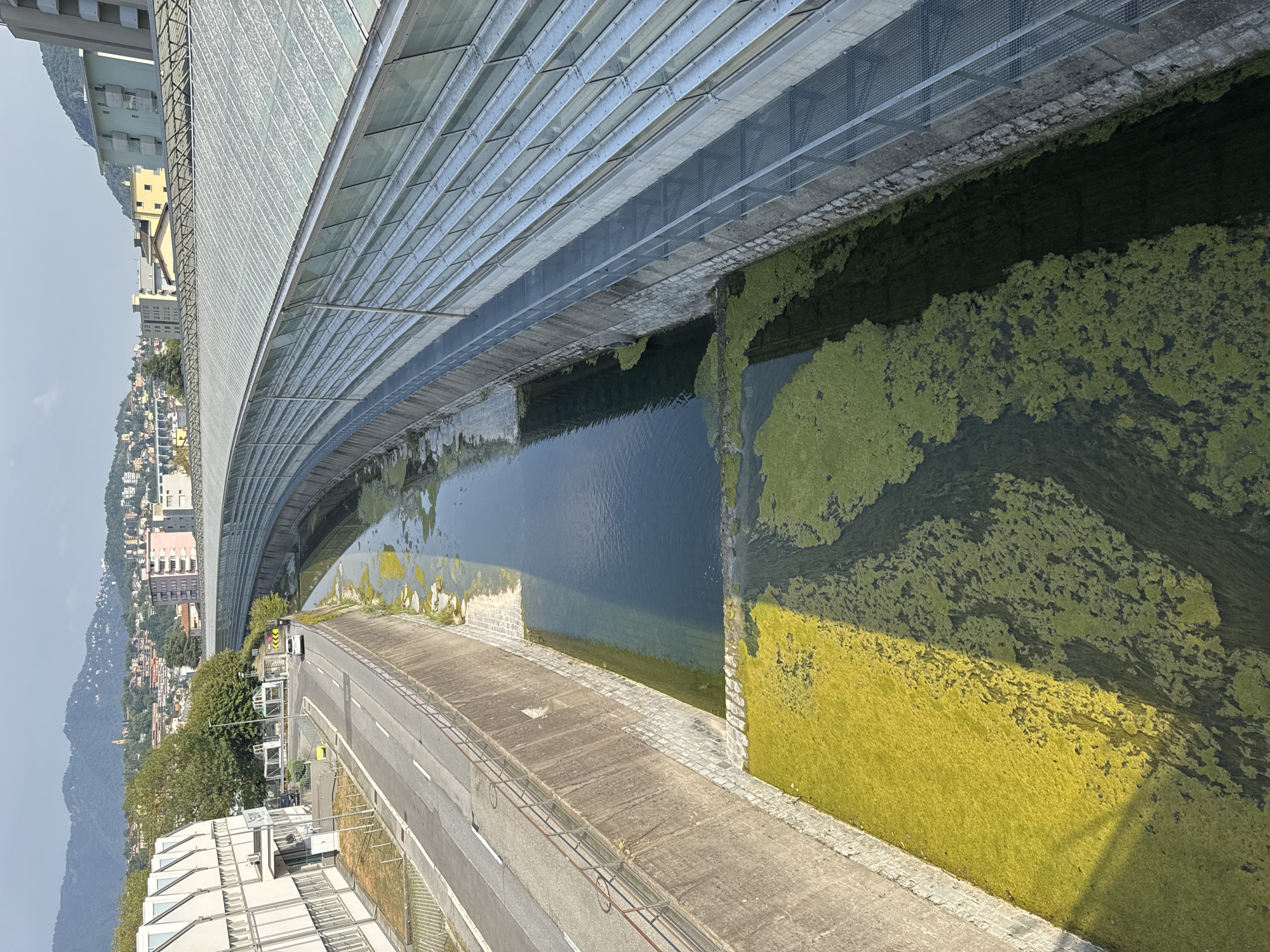 The image shows a view looking down onto a canal in Morbio Inferiore, Switzerland. The canal is lined with concrete walls and has a metal grate walkway along its right side. The water in the canal is partially covered with green algae. To the left of the canal is a road with a white car driving on it. Beyond the road, there is a modern building with a white facade and slanted windows. In the background, there are hills with buildings and trees, indicative of a town or city. The sky is clear and blue.