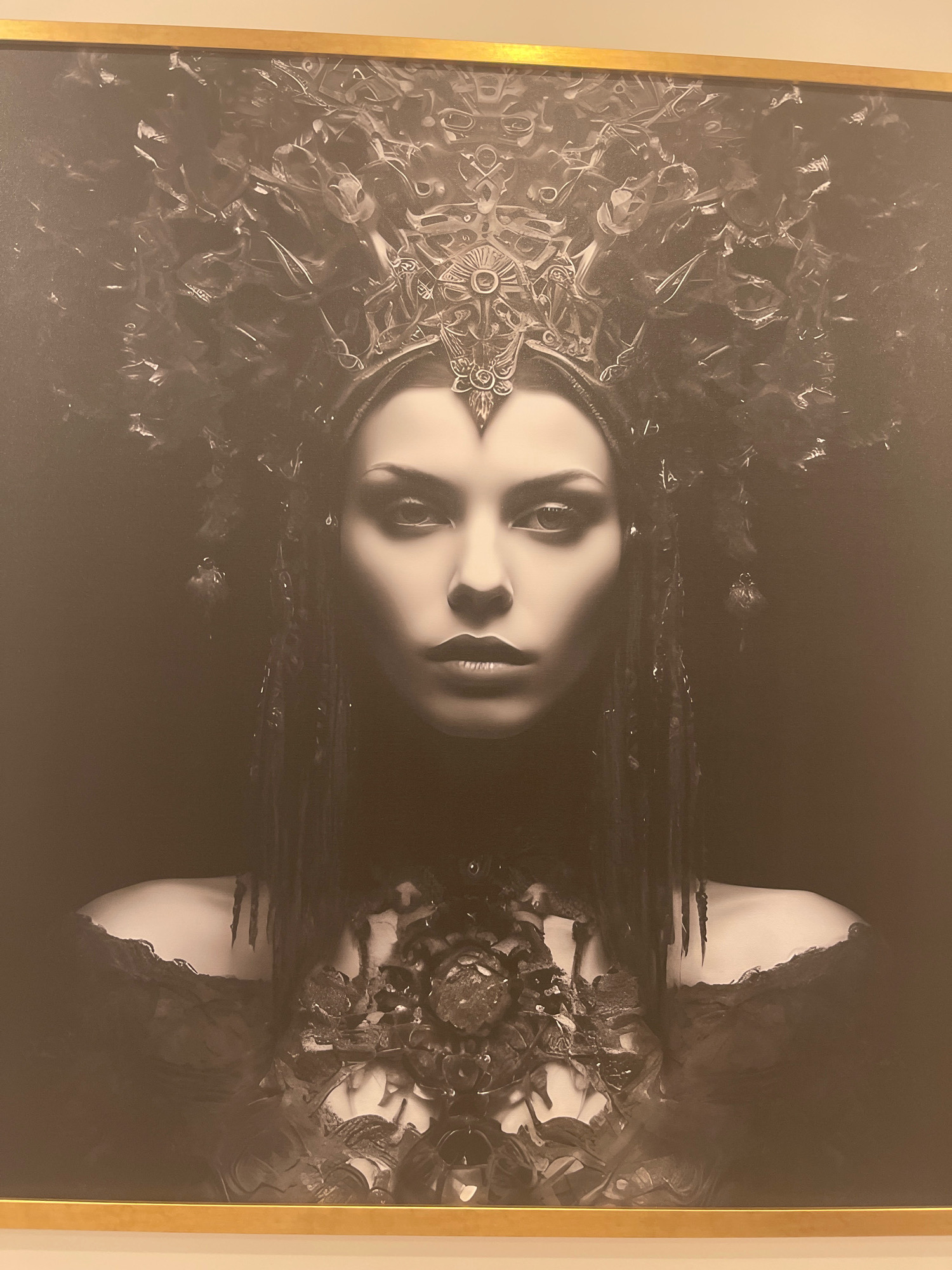 The image is a framed black and white portrait of a woman wearing an elaborate headdress and ornate jewelry. The woman's face is centrally positioned and illuminated, with striking eyes and dark lips. Her headdress is complex and appears to be made of various organic and metallic elements, extending upwards and outwards. She also wears a heavily embellished collar or chest piece. The background is dark and fades to black, creating a dramatic contrast with the subject. The style of the artwork suggests it is likely a digital creation or a heavily manipulated photograph. The image is a photograph of this artwork hanging on a wall, with a thin gold frame visible around the edges. The location of this artwork is unknown and cannot be determined from the image provided, and therefore its location in Moscow, Russia, is not ascertainable.