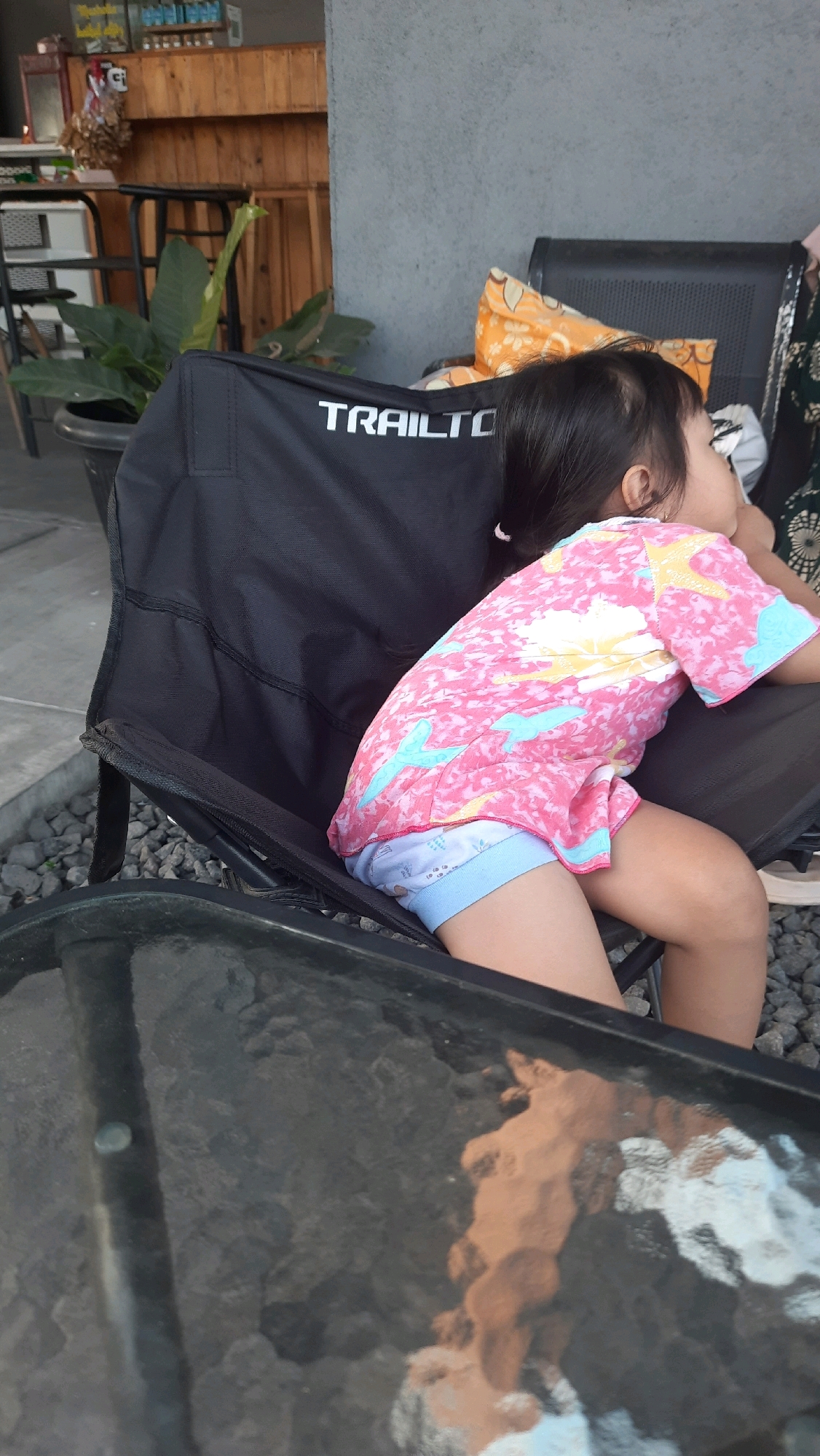 A young child is seen from the side, seated in a black camping chair in what appears to be an outdoor cafe in Jajag, Indonesia. The child is wearing a pink t-shirt with a colorful starfish and wave pattern, and light blue shorts. The child's head is turned to the right, with their profile visible. Behind the chair, an orange pillow with a floral pattern is visible. In the background, there's a wooden counter with shelves, plants in pots, and a concrete wall. The foreground shows a reflective surface, possibly a glass table, with distorted reflections. The ground is covered in small grey gravel.