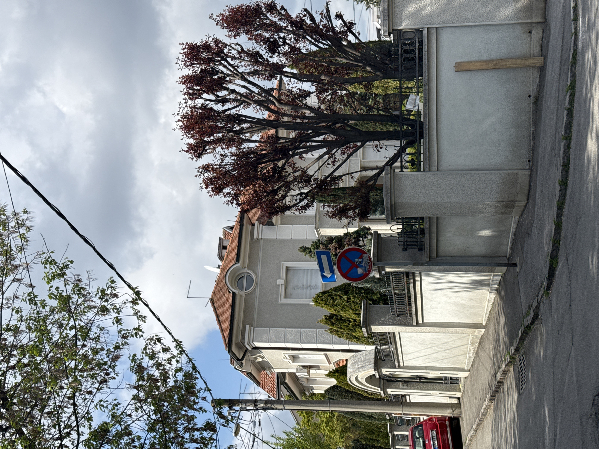 The image shows a residential street in Vozdovac Urban Municipality, Serbia. The buildings are light-colored with red-tiled roofs and architectural details. A tree with reddish-brown leaves partially obscures one of the buildings. A street sign with a blue arrow pointing down and a red circle with a crossed-out symbol is visible. The sidewalk is concrete, and the street is asphalt. A red vehicle is partially visible at the bottom of the frame. Overhead power lines run across the top left of the image. The sky is cloudy.