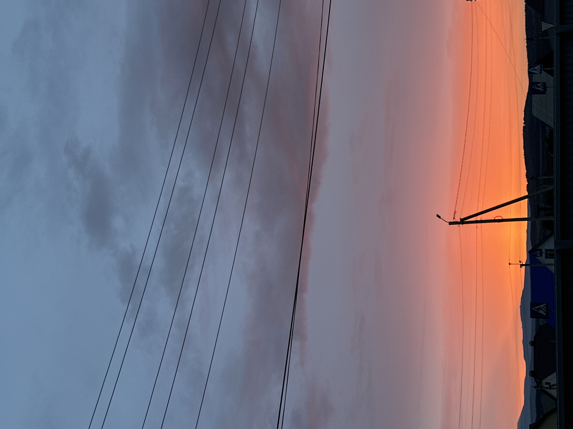 A landscape photograph captures a dramatic sunset or sunrise over a rural or semi-urban setting. The sky occupies the majority of the frame, showcasing a striking transition from dark, cloudy grey on the left to vibrant shades of orange, pink, and yellow on the right.
Several dark, thin utility wires cut diagonally across the upper portion of the sky, running from the top-left towards the lower-right. On the right side, a tall utility pole with a single streetlight fixture is silhouetted against the brightly colored sky, with more wires extending from it.
Below the colorful horizon, a dark silhouette of hills or mountains stretches across the right half of the image, suggesting a rugged backdrop. At the base of these hills, the dark outlines of buildings or houses indicate a small settlement. Two triangular road signs are partially visible near the top-right of the foreground structures; one clearly displays a symbol of two figures walking, characteristic of a pedestrian crossing sign.
The time of day is unequivocally dawn or dusk, with the rich, warm colors of the sky strongly suggesting a sunset. The weather appears partly cloudy, with heavier, darker clouds on the left contrasting with the clearer, vibrant sky on the right. No discernible human activity or explicit text is visible, beyond the symbols on the road signs.