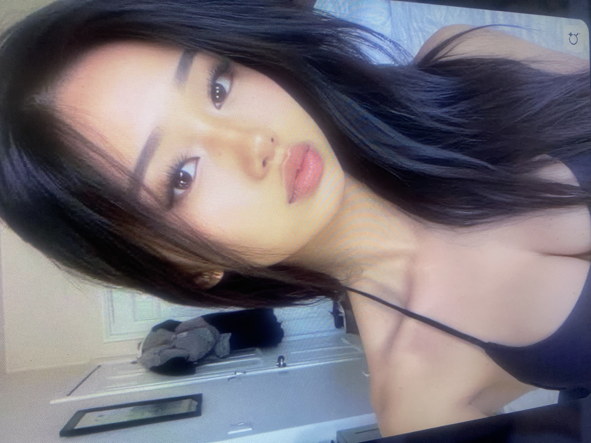 The image is a close-up portrait of a young East Asian woman with long, dark hair. She is wearing a black bikini top and looking towards the camera. Her makeup includes defined eyebrows, eyeliner, and lip gloss. The background appears to be an indoor setting, possibly a bedroom, with a closet door, a framed picture on the wall, and a backpack hanging on the door. The lighting is soft, and the image has a slightly grainy quality. There are no recognizable landmarks or clues that would place this image in Samara, Russia.