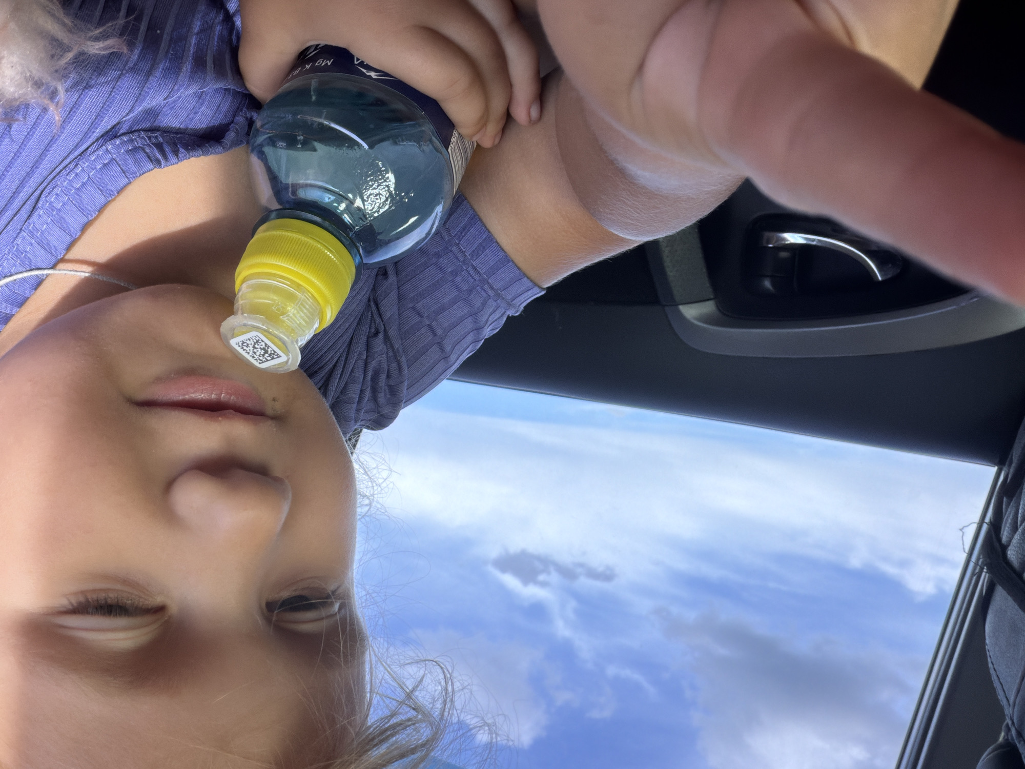 A close-up, low-angle shot captures a young child in the foreground, holding a bottle of water. The child, with light-colored hair and fair skin, is looking down, slightly to the right, with one eye squinted. They are wearing a purple, ribbed shirt. The water bottle has a yellow cap and a clear plastic seal with a QR code. A white string necklace is visible around the child's neck. The background shows a portion of a car's interior, including a dark door panel with a chrome handle, and a bright blue sky with scattered white clouds, suggesting the car is in motion. The image was taken in Novoe Mavrino, Russia.