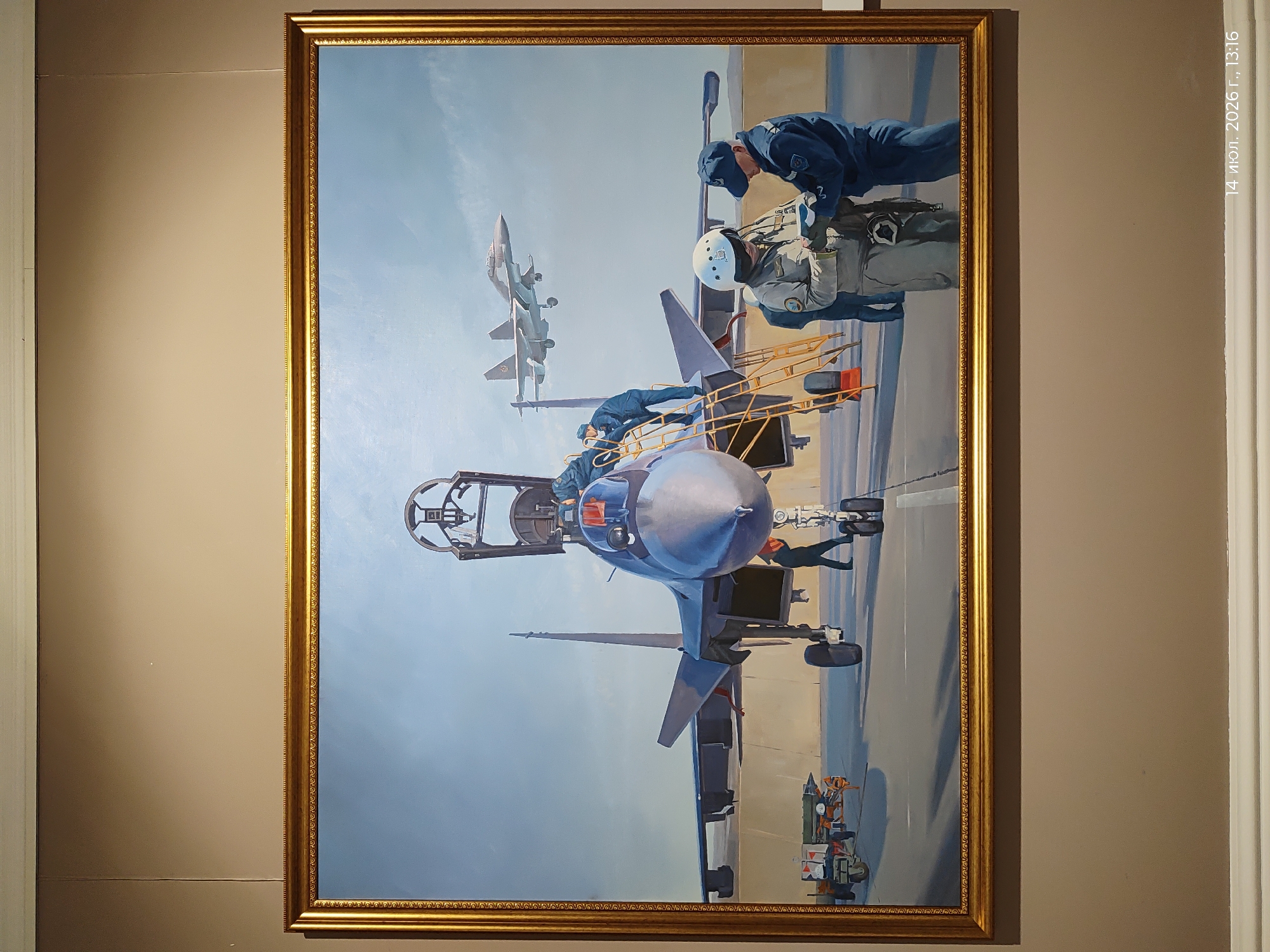 A realistic oil painting in a gilded frame depicts a scene on an airfield with a fighter jet in the foreground. The jet, with its canopy open, is being attended to by several figures in military uniforms. Two figures in flight suits and helmets are standing in front of the jet, appearing to consult documents. Another figure in a dark blue uniform stands beside them, also looking at the documents. Several other figures are visible near the jet and on a yellow ladder attached to its side. In the background, another fighter jet is in flight against a pale blue sky. To the left of the main jet, a trailer with what appears to be munitions is parked. The ground is paved, indicating an airport or military base. The overall impression is one of preparation and readiness for flight operations. The watermark at the bottom right of the image reads "14 июл. 2026 г., 13:16", suggesting the image was taken on July 14, 2026, at 13:16. There is no specific indication of the location being the city of Astana, Kazakhstan, within the painting itself or the visible elements of the image.