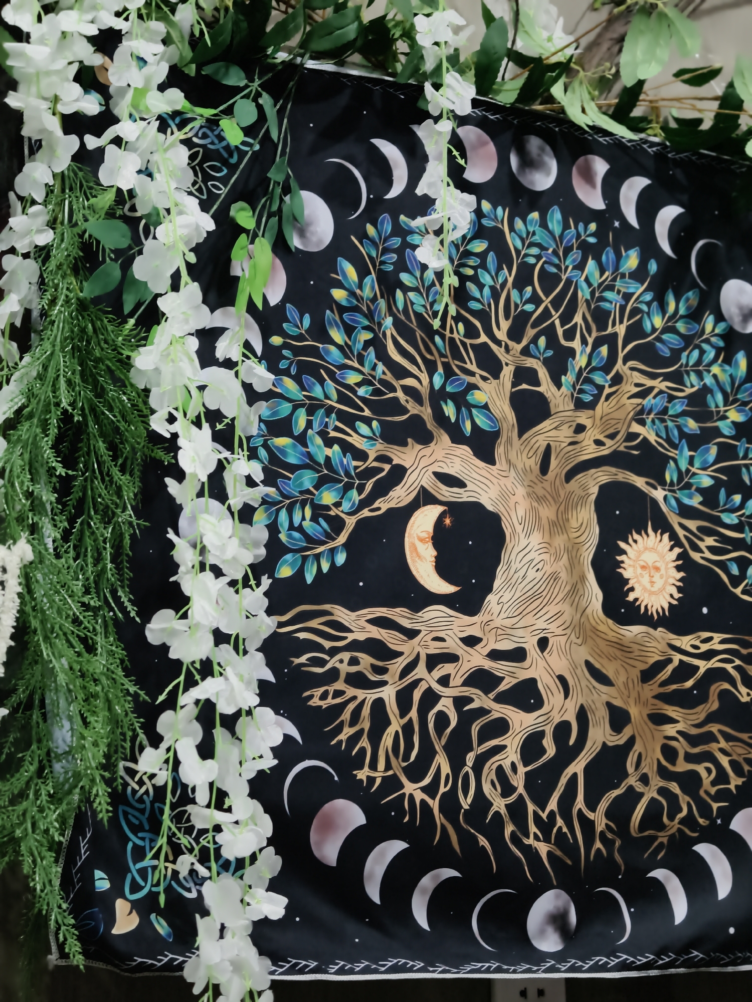 The image displays a tapestry with a Tree of Life design hanging against a dark background. The tree has golden-brown trunk and branches, adorned with blue and green leaves. The tapestry also features celestial elements, including a crescent moon with a face and a sun with a face, as well as phases of the moon arranged in a circular pattern around the tree. White artificial flowers and green artificial foliage hang from above, partially obscuring the tapestry. The bottom edge of the tapestry has a decorative border. The scene does not provide any specific information about its location within the city of Đà Nẵng, Vietnam. There are no people or actions depicted in the image.