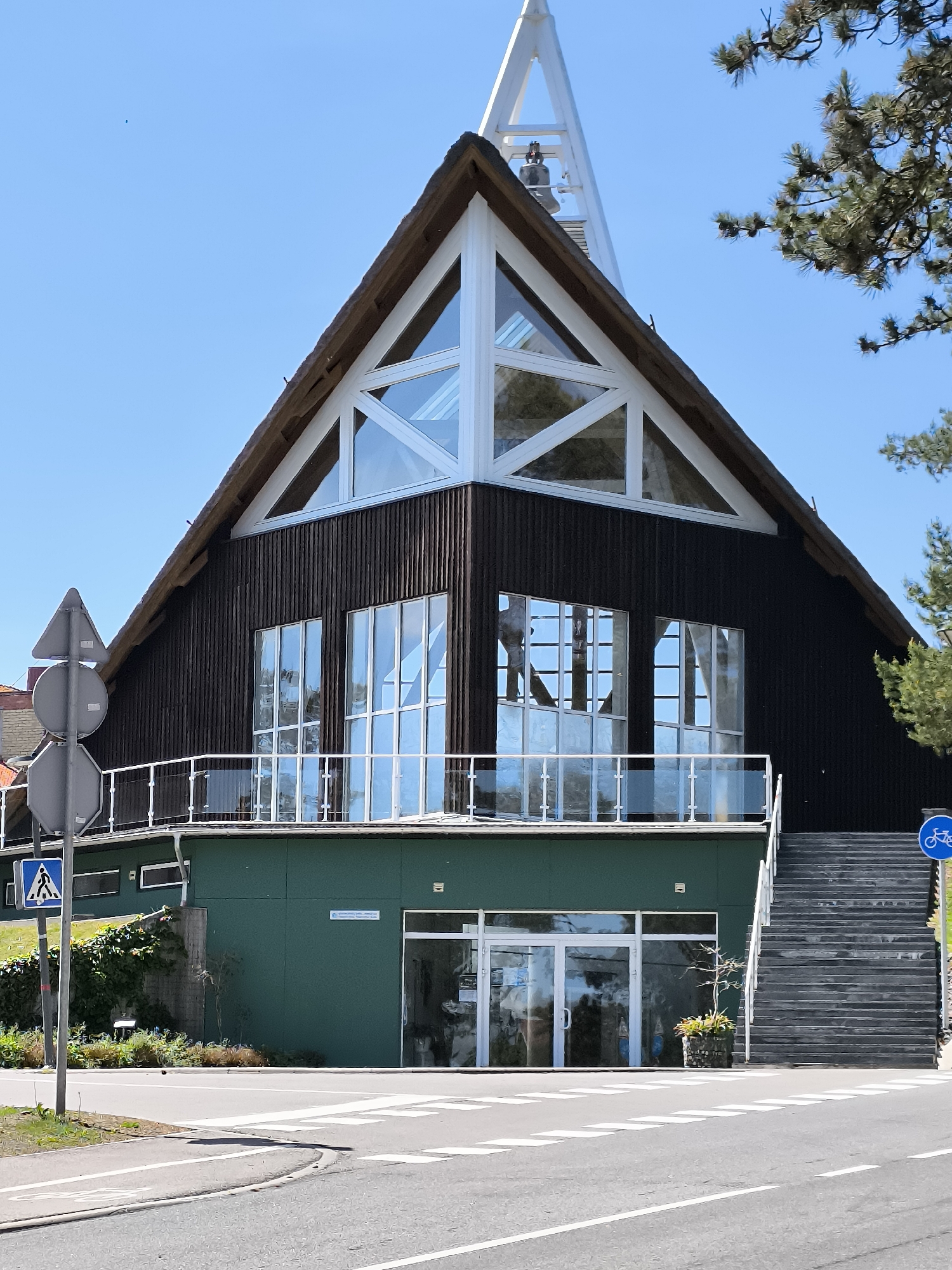 The image depicts the front of a modern building with a distinctive A-frame roof structure, likely made of wood or thatch, with a prominent white triangular window. The upper facade is clad in dark, vertical wooden planks, and features several large, multi-paned windows with reflective glass. A white railing runs along a balcony that spans the width of this upper section. Below this, the building has a green-colored lower level with glass double doors leading to the entrance. To the right of the entrance, a long, outdoor staircase with dark treads and white railings ascends along the side of the building. A blue circular sign with a white bicycle icon is visible at the top of the stairs. A pedestrian crossing is marked on the asphalt road in front of the building, with a blue square sign displaying a white pedestrian symbol on the left side of the image. The sky is clear and blue, and a tree with green foliage is visible on the right side. The building is situated in a location that appears to be a public area, possibly near a park or a coastal region, given the clear sky and vegetation. This building is likely the Klaipėda Sea Museum's exhibition pavilion located in Nida, Neringa, Lithuania.