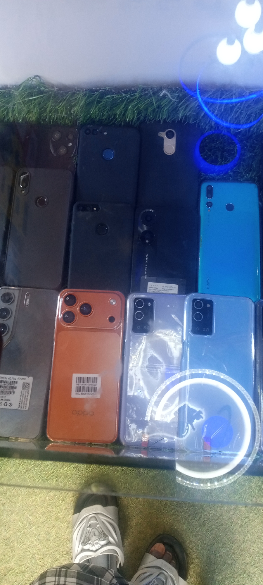 The image depicts a collection of used smartphones arranged on a display counter, likely for sale. The counter is covered with artificial turf. A glass surface separates the phones from the viewer.
The phones vary in color and design, suggesting a mix of models and brands. Some are in protective cases. Price tags or IMEI labels are visible on a few of the phones.
A partial reflection of a person is visible in the glass, showing feet wearing sandals and plaid patterned clothing. There is a bright, ring-shaped light source reflected in the glass.
The precise location within Jos, Nigeria cannot be determined from the image.