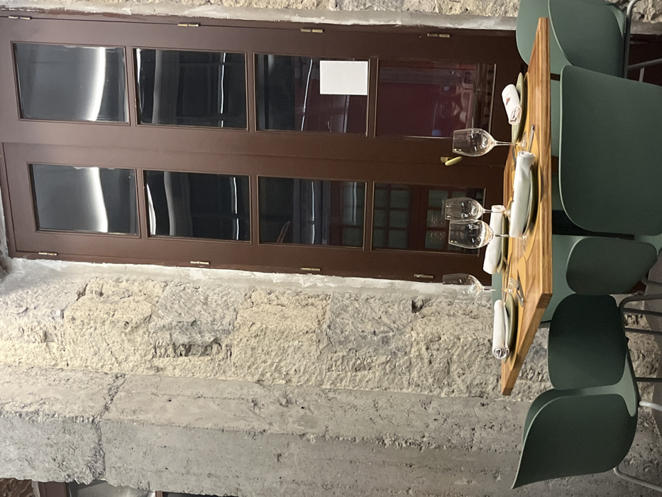 The scene shows an outdoor dining area located on the patio of a restaurant in Las Palmas de Gran Canaria, Spain, with a view of the nearby street. The patio is enclosed on three sides by a concrete wall with a large brown wooden door on one side and a large concrete pillar with chipped paint on the other. There are five green chairs around a table, two of which are occupied, and one of which is empty. The table has a wooden top and a metal base. On the table are four place settings with white napkins, silverware, and glasses. The table has a glass with a red liquid on it. There are four wine glasses, one of which is on the empty setting, and three of which are filled. The table is decorated with a wooden tray with a pattern of small, alternating light and dark squares. Two of the glasses on the table have a red liquid in them. The door appears to be made of two panels and has a brass handle and several hinges. The door is closed and the glass panels are slightly frosted. There are several other objects visible in the reflection in the glass door panels, but their details are not clear. The scene is lit by the sun shining through the glass panels of the door. The air temperature appears to be moderate. The image was taken on a clear day with minimal cloud cover.