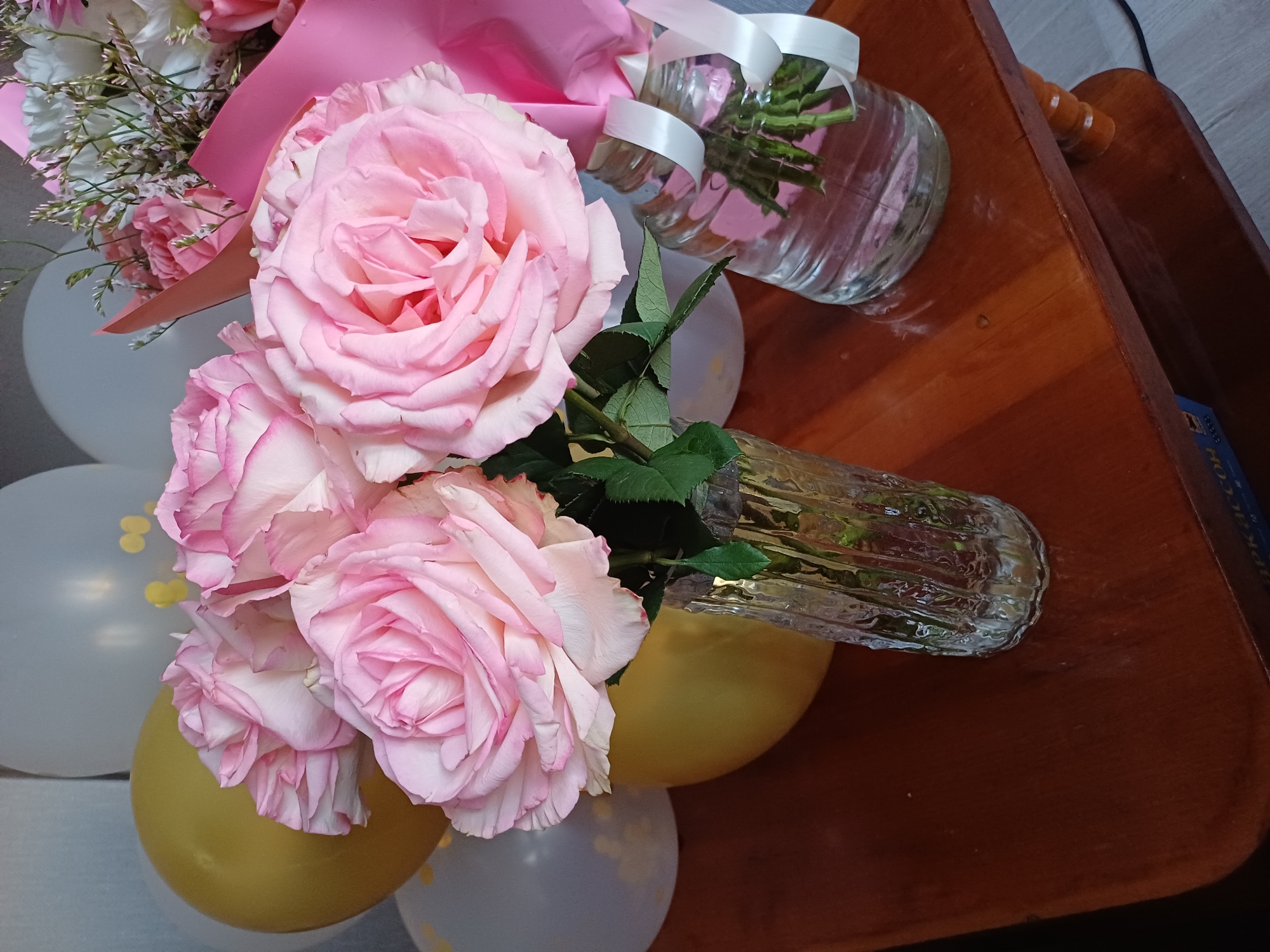 A close-up shot shows a bouquet of pink roses, interspersed with white carnations and other filler flowers, wrapped in pink paper and tied with white ribbon. The bouquet is placed in a clear glass vase that appears to be partially filled with water. The vase is resting on a polished wooden surface, possibly a table. In the background, several balloons, some white and some a golden yellow, are visible. One of the balloons is partially obscured by the bouquet and vase. The lighting suggests it may be indoors during the day. The overall impression is that of a decorative arrangement, possibly for a celebration or gift. There is no visible text in the image.