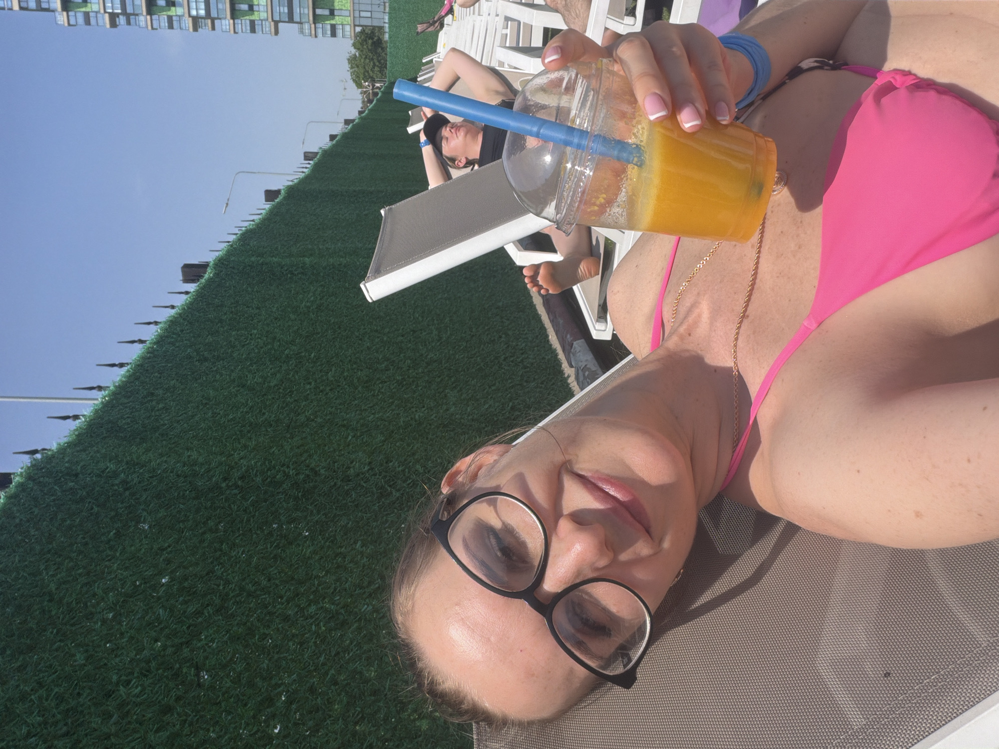 A woman in a pink bikini and black eyeglasses is lying on a beach chair, holding a plastic cup with orange liquid and a blue straw. Another woman is sunbathing in the background, also on a beach chair. A green fence separates the sunbathing area from the cityscape, which shows modern apartment buildings. The photo was taken in Moscow, Russia.