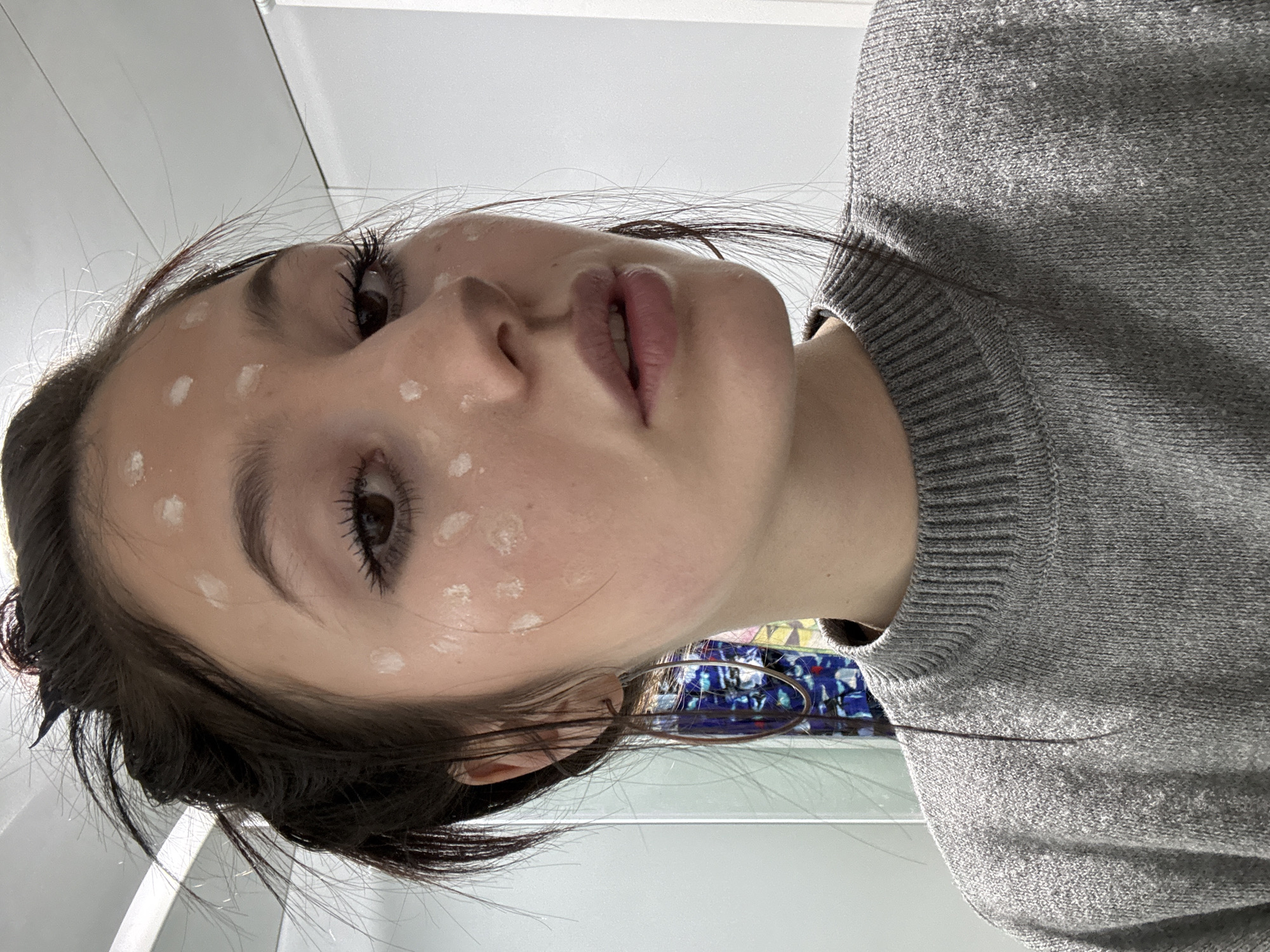The image presents a close-up, angled portrait of a young person, likely female, with dark hair, looking upwards and slightly to the right of the camera. Her face features numerous small, circular white applications, indicative of skincare product or foundation dots being applied. She has prominent dark eyebrows and long eyelashes. Her lips are naturally colored and slightly parted.
She is wearing a grey knitted sweater with visible ribbing around the neckline and cuff. A large, thin, metallic hoop earring is visible on her left ear. The background is predominantly bright white, suggesting an indoor setting with strong illumination. A hint of vibrant, multi-colored patterns, possibly a mosaic or abstract design, is visible in the blurred background behind her left ear. The person's expression appears neutral and composed. No visible text is present in the image. The location context provided is Фоки, Russia.