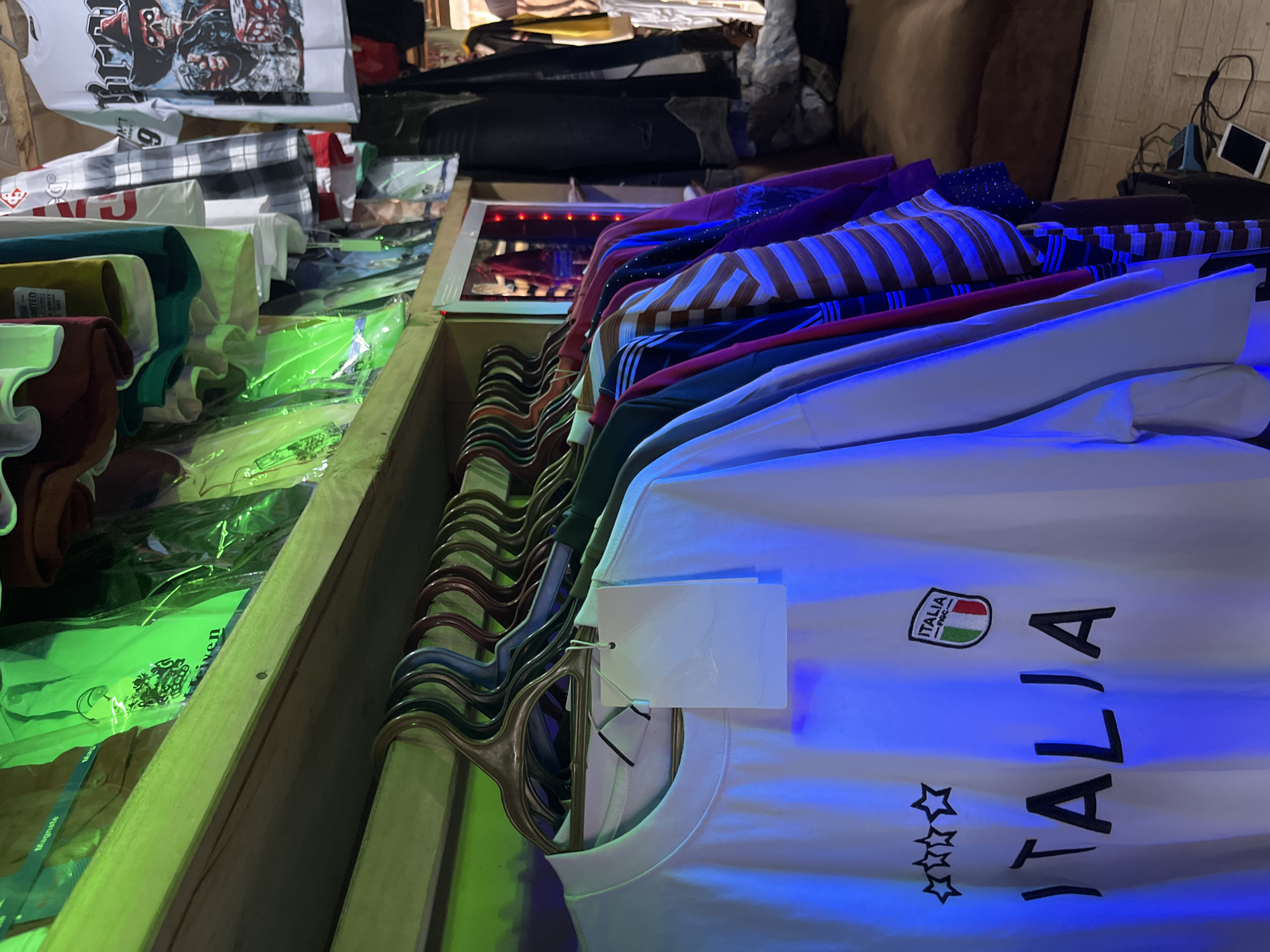 The image depicts a clothing store in Bukuru, Nigeria. The foreground is dominated by a white t-shirt with black lettering that reads "ITALIA" and a crest with the word "FIGC" and the Italian flag. The t-shirt is hanging on a hanger on a clothing rack, illuminated by blue and green lights. Behind this t-shirt, other clothing items are visible, including shirts with stripes and various colors, also hanging on hangers. In the background, shelves filled with more clothing are visible, some in plastic packaging, illuminated by green light. A wooden shelf runs across the top of the image. The environment appears to be an indoor retail space, possibly a stall or small shop, with wooden structures and a dark background suggesting a cluttered storage area. There are no people visible in the image.
