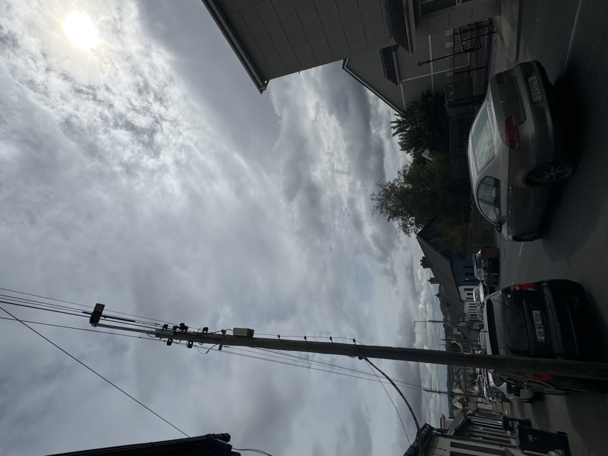 The image depicts a street scene in Waterford, Ireland, under an overcast sky with the sun breaking through clouds in the upper right. A wooden utility pole with numerous wires and a box attached to it stands prominently on the left side of the road. The street is lined with houses and parked cars. In the foreground, a black hatchback with license plate 08-WX-1231 is parked on the left. Further down the street, other vehicles are parked on both sides, including a silver sedan that appears to be in motion. The architecture of the houses suggests a residential area. The overall atmosphere is subdued due to the cloudy weather, with the sun creating a brighter area in the sky.