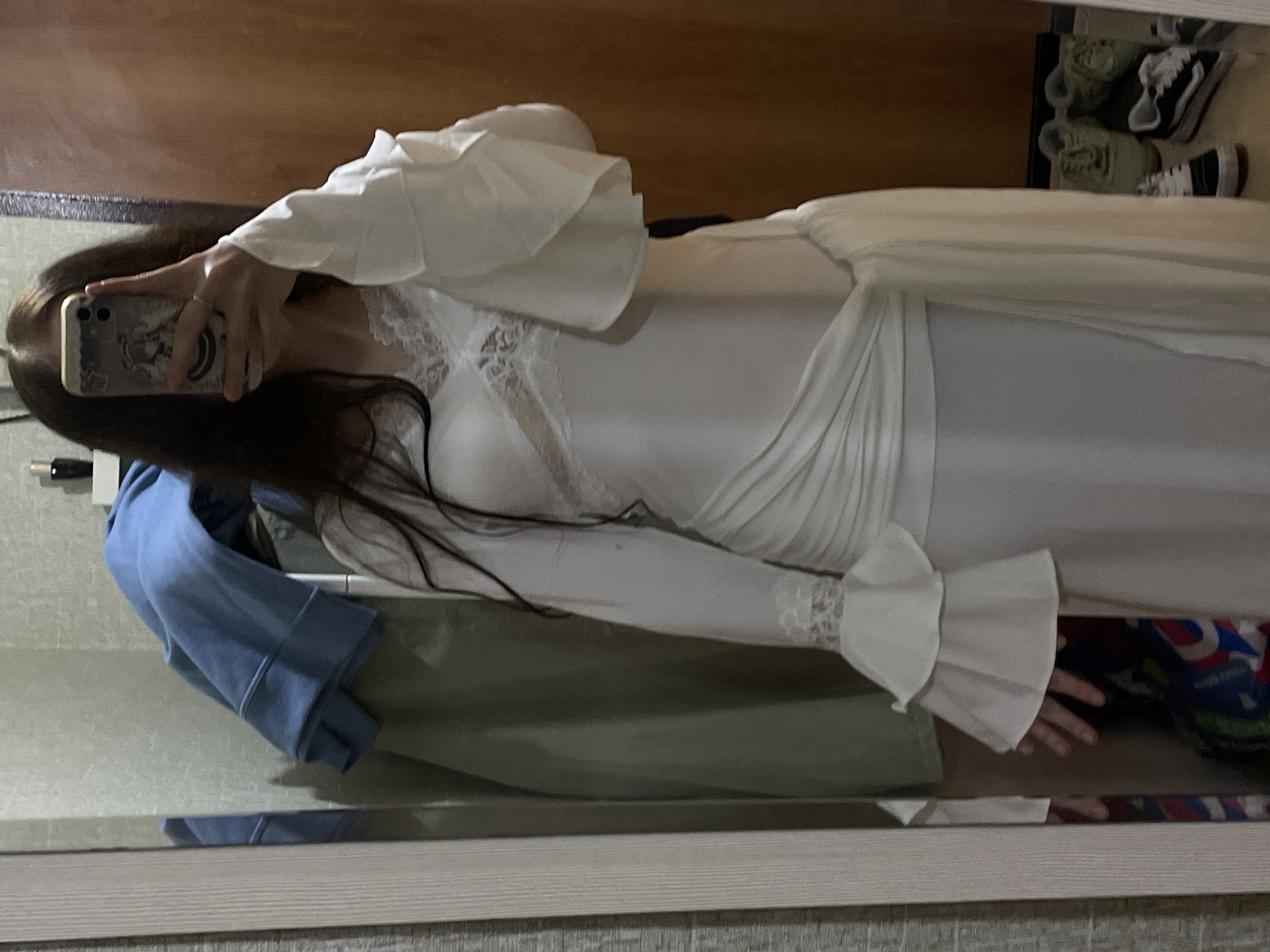 The image shows a person, likely a woman given the dress and long hair, holding a phone to take a mirror selfie. The person is wearing a long white dress with lace detailing at the bust and ruffled bell sleeves. The dress appears to be made of a sheer or light fabric. In the reflection of the mirror, a blue hoodie is visible, draped over something out of frame. Behind the person, on a shelf or surface, a pair of Vans sneakers are visible, suggesting a casual indoor setting. The background appears to be a room with wooden furniture and textured walls. The lighting is dim, suggesting it might be evening or an indoor setting with low light. There is no visible text in the image. The overall scene conveys a sense of personal space and perhaps an attempt to capture a moment or outfit.