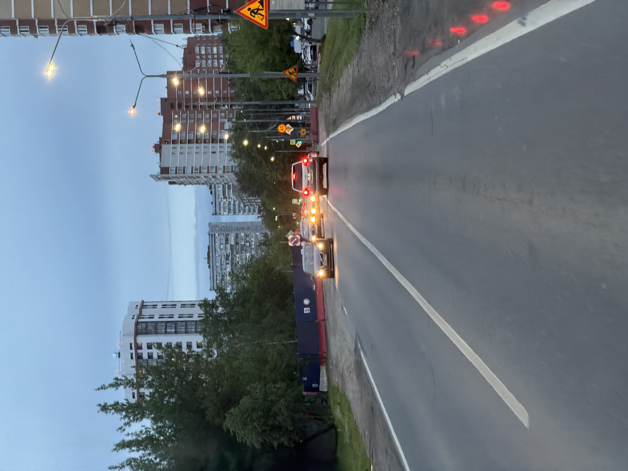 The image captures a street scene in a city with multi-story residential buildings and streetlights lining the road. The road surface appears wet, possibly after rain. There are several vehicles visible on the road, including cars and a van, with their headlights and taillights illuminated. Traffic signs and construction warnings are also present. The environment suggests an urban setting, potentially during early evening or late afternoon, given the ambient light. The image does not provide sufficient detail to determine the precise location within the city or to identify any specific people.
