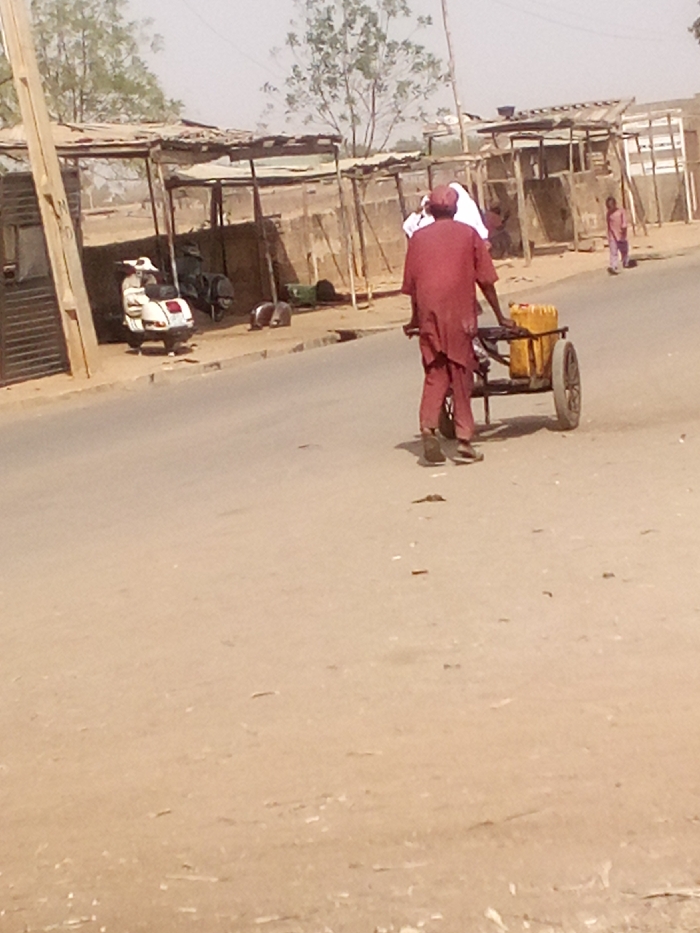 The image displays a daylight street scene in Tudun Murtala, Nigeria. A male figure, viewed from behind, wears a maroon long-sleeved tunic and matching trousers, and is pulling a two-wheeled handcart loaded with two yellow plastic jerrycans. Immediately behind this individual, a second person in white attire is partially visible. The foreground consists of a light brown dirt road surface with a grey paved section. To the left, a white vintage-style scooter is parked on the dirt shoulder adjacent to a dark, possibly corrugated metal gate and a light-colored wooden pole. Further into the mid-ground, a series of rudimentary stalls or shelters with corrugated metal or tarpaulin roofs, supported by wooden posts, line the street. A light-colored wall stands behind these structures. In the far right background, a third individual in a light pink garment walks away from the viewer. Sparse green trees are visible in the background against a light sky.
