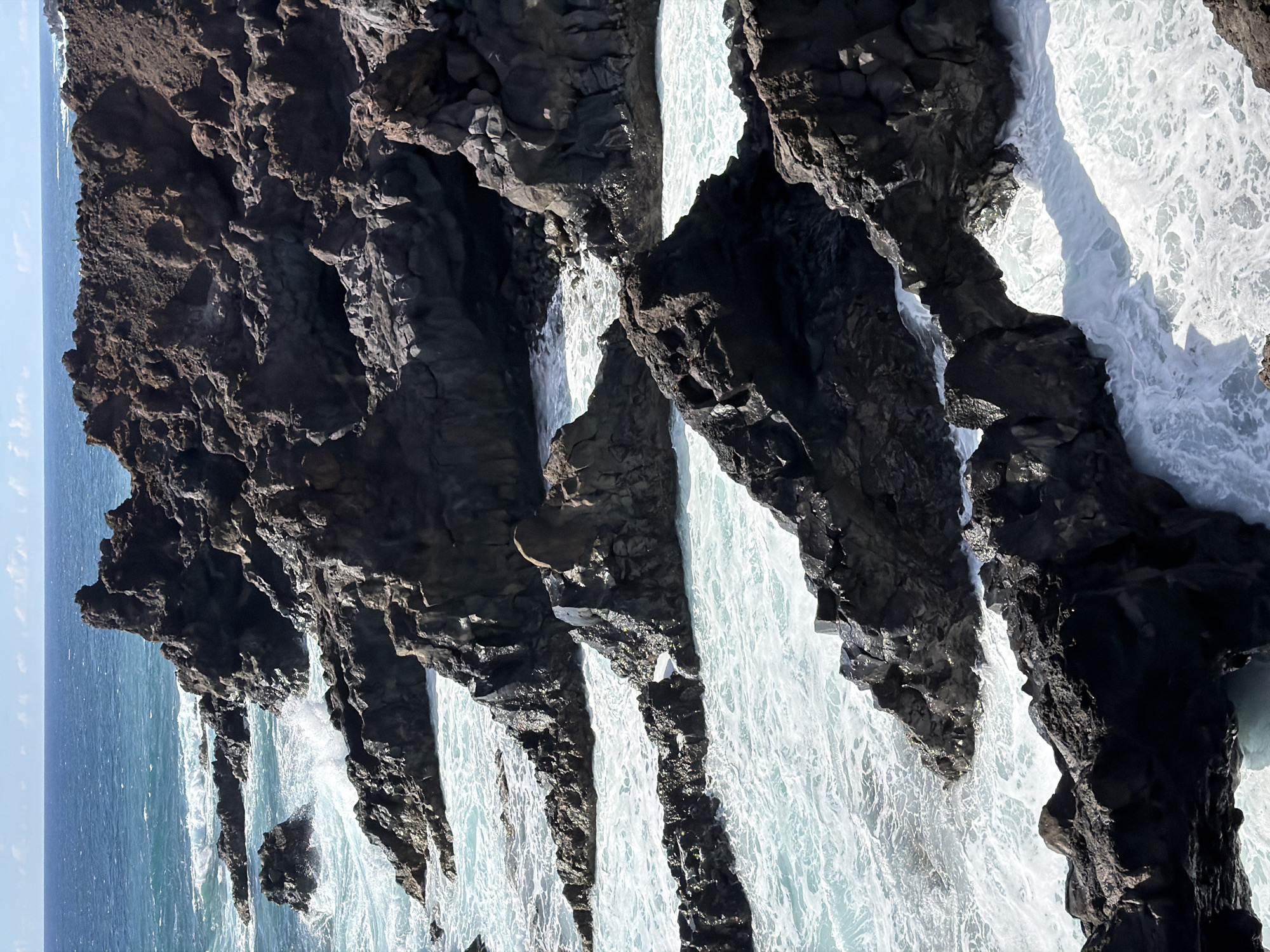 This photograph captures a scene of rugged, dark volcanic rock formations meeting turbulent ocean waters. White foamy waves surge through narrow channels and around rocky outcrops, creating a dynamic and powerful visual. The sky, a pale blue, is barely visible at the top left, suggesting the scene is focused on the interaction between the land and sea.
The location is unidentified.