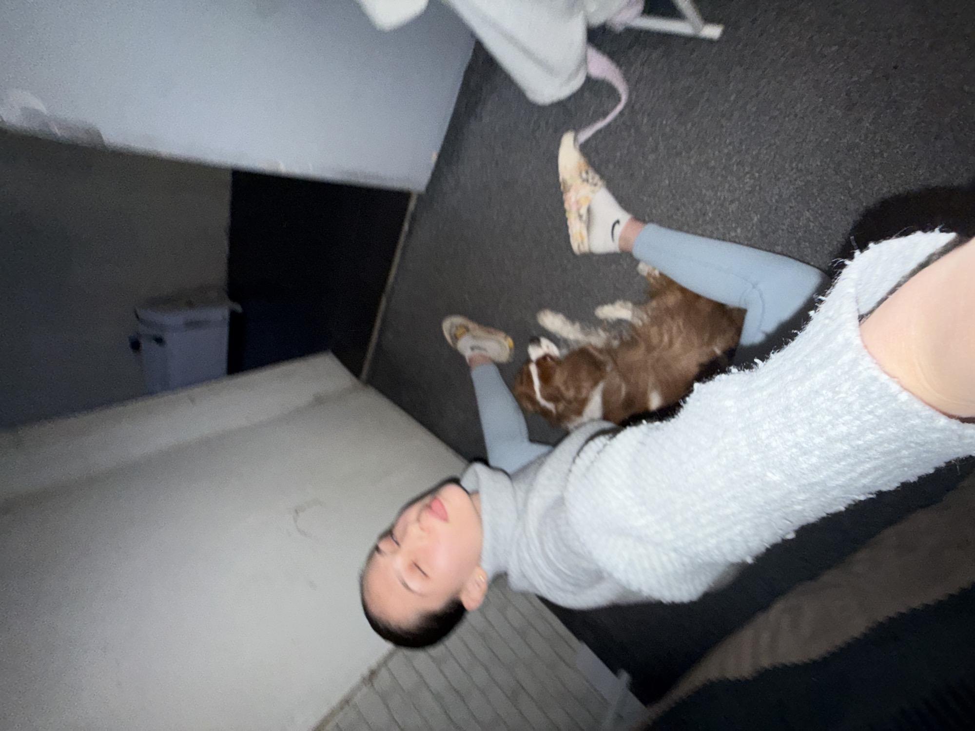An angled, high-perspective indoor photograph captures a person and a dog resting on a dark, textured floor. The person, appearing to be a young woman, lies on her side, wearing a light grey or off-white turtleneck sweater and light blue pants. Her hair is pulled back, and her eyes are closed, with lips slightly parted, suggesting she is sleeping or deeply relaxed. She wears white socks with a black logo, possibly Nike, and light-colored slip-on shoes with a subtle pattern.
Beside her, a brown and white dog also lies down, appearing to be resting comfortably. The floor is a dark, possibly asphalt or industrial-style textured surface. In the background to the left, a large light grey or white horizontal surface, possibly a table or bench, is visible. Further back, a white cooler or storage box is discernible against a darker wall. A light-colored strap, possibly from a bag, is near the person's feet. The lighting is dim, suggesting an evening or indoor setting with artificial light. The overall mood is calm and peaceful. The scene takes place in Sofia, Bulgaria.