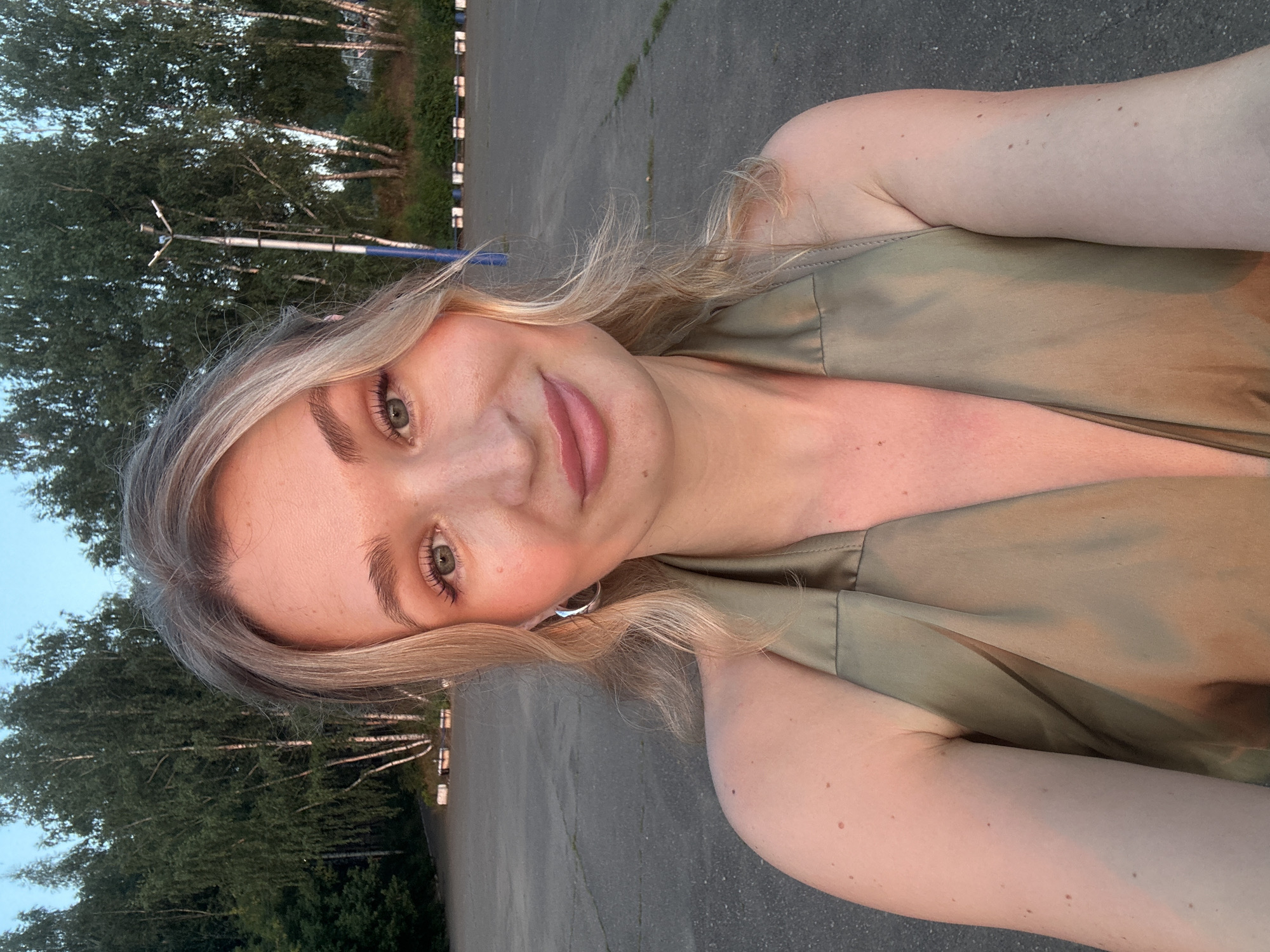 A young, blonde woman with light skin and light green eyes is taking a selfie outdoors. She is wearing a light olive green satin halter top. She has large hoop earrings. Her hair is styled in soft waves around her face. Her lips are slightly parted, and she has a subtle smile. The background consists of a paved area with some trees and a streetlamp. The lighting suggests it is late afternoon or early evening. The location is the parking lot of the "Energetik" Sports Complex in Chaikovsky, Russia.
