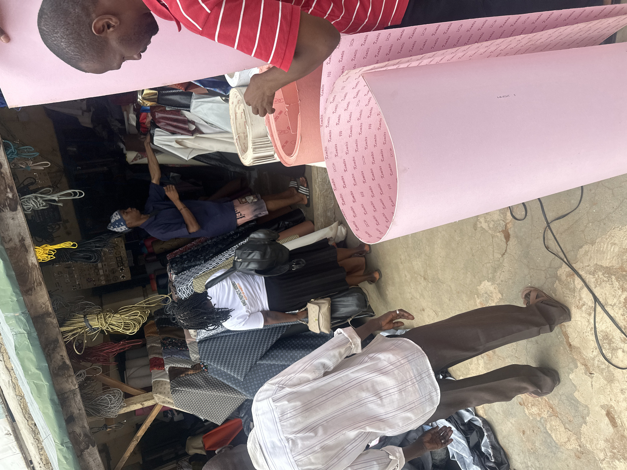 This image depicts an active, open-front market stall or shop located in Jos, Nigeria, specializing in various materials. In the foreground to the right, a dark-skinned man wearing a red and white striped polo shirt is handling a large roll of light pink foam sheet, which is distinctly marked with repeating "Kemter", "910", and "DOD" text. To the left, a second dark-skinned man is observed from the back, dressed in a white long-sleeved shirt with subtle vertical stripes, brown trousers, and brown sandals; a ring is visible on his right hand.
In the mid-ground, a dark-skinned woman with braided hair, also seen from the back, wears a white t-shirt with the legible inscription "KINGS PROPHETIC ASSEMBLY CITY OF JOS", a black pleated skirt, and carries both a black backpack and a light brown handbag. Further inside the shop, to the left, a third dark-skinned man in a blue t-shirt and a blue and white patterned cap reaches into a display of various rolled materials.
The shop interior is densely arranged with numerous rolls and bolts of patterned fabrics, including dark grey geometric designs and black and white patterns. Various colored leather or vinyl sheets in white, red, brown, gold, and black are also stacked. Coiled bundles of yellow, red, grey, and black cords hang from wooden overhead beams, which support a green corrugated metal sheet forming an overhang above the entrance. A small display of metallic items is visible on the far right within the shop. The ground is an uneven, light-colored concrete or packed earth surface, across which a black electrical cord extends.