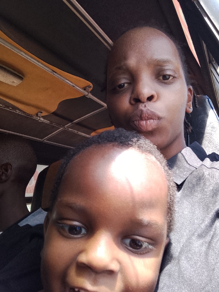 A woman and child are in a vehicle, possibly a bus or van. The woman is behind the child, who is close to the camera. The woman has dark skin, short hair, and is wearing a gray top. She is pouting her lips toward the camera. The child has darker skin and is smiling, revealing teeth. The vehicle interior has a yellow-tanned fabric lining with metal supports. Part of another person's head is visible to the left.