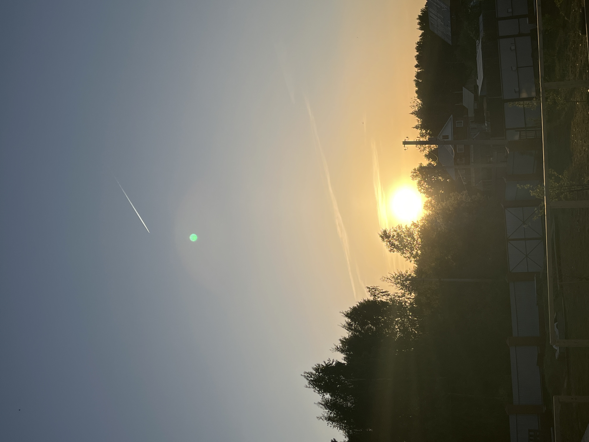 The image shows a sunset in a rural or semi-rural area, likely in Chaikovsky, Russia, due to the mention of the city. The sky transitions from blue to orange as the sun sets behind trees and buildings. A contrail is visible in the blue portion of the sky. Silhouetted trees and structures suggest residential or agricultural outbuildings. The image is taken from a low angle, possibly from ground level, looking towards the setting sun. There are no people visible.