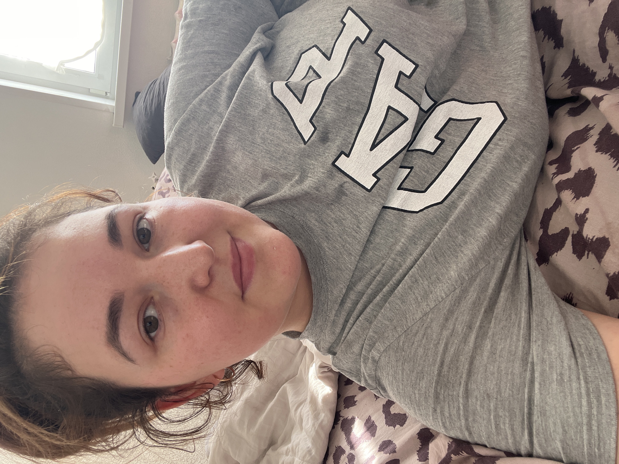 This media file is a selfie taken by a young person lying on a bed. The subject is wearing a grey sweatshirt with the word "GAP" in large white letters with black outlines across the chest. They have brown hair, brown eyes, and fair skin with some freckles. Their expression is relaxed and neutral. The bedsheets are white, and there is a patterned pillow with a brown and cream animal print visible. To the left of the frame, a window with a white frame and a sheer white curtain is visible, letting in natural light. The overall scene suggests a casual, indoor setting, likely a bedroom, during the daytime. There are no specific cues to indicate the weather, but the natural light suggests it is not dark outside. The location is Chelyabinsk, Russia.