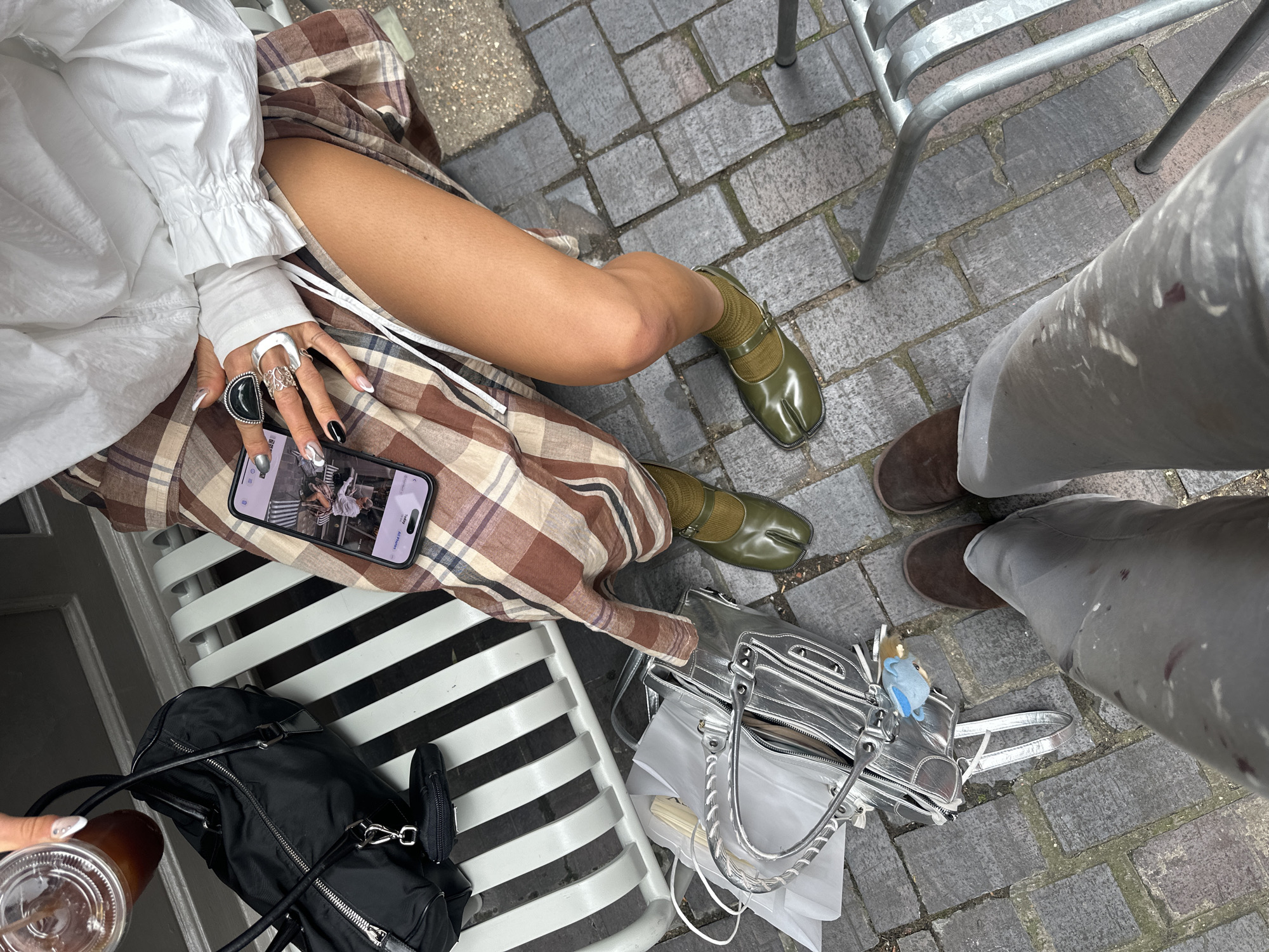 An eye-level, high-angle shot depicts two individuals seated outside in what is suggested to be London, United Kingdom, evidenced by the paving style and the presence of gray painted iron furniture.
The subject on the left, a lighter-skinned person, is partially visible. They are wearing a voluminous white long-sleeved shirt with ruffled edges at the wrist, and a tan and brown plaid skirt that covers the top of their legs. The person is wearing green split-toe shoes and matching socks. They are holding a phone in their left hand, which is adorned with silver rings and unique manicure styles of white, black, and silver. The phone displays a picture in the "All Photos" section.
On the lower left is a black zippered backpack with silver hardware, and a partially visible cup with a straw, likely containing a beverage. A silver handbag is placed between the two people.
To the right, another individual is partially visible from the knees down. They are wearing gray pants and brown suede shoes. The legs are positioned near a gray iron chair.
The setting is a brick-paved area, possibly a patio or sidewalk.