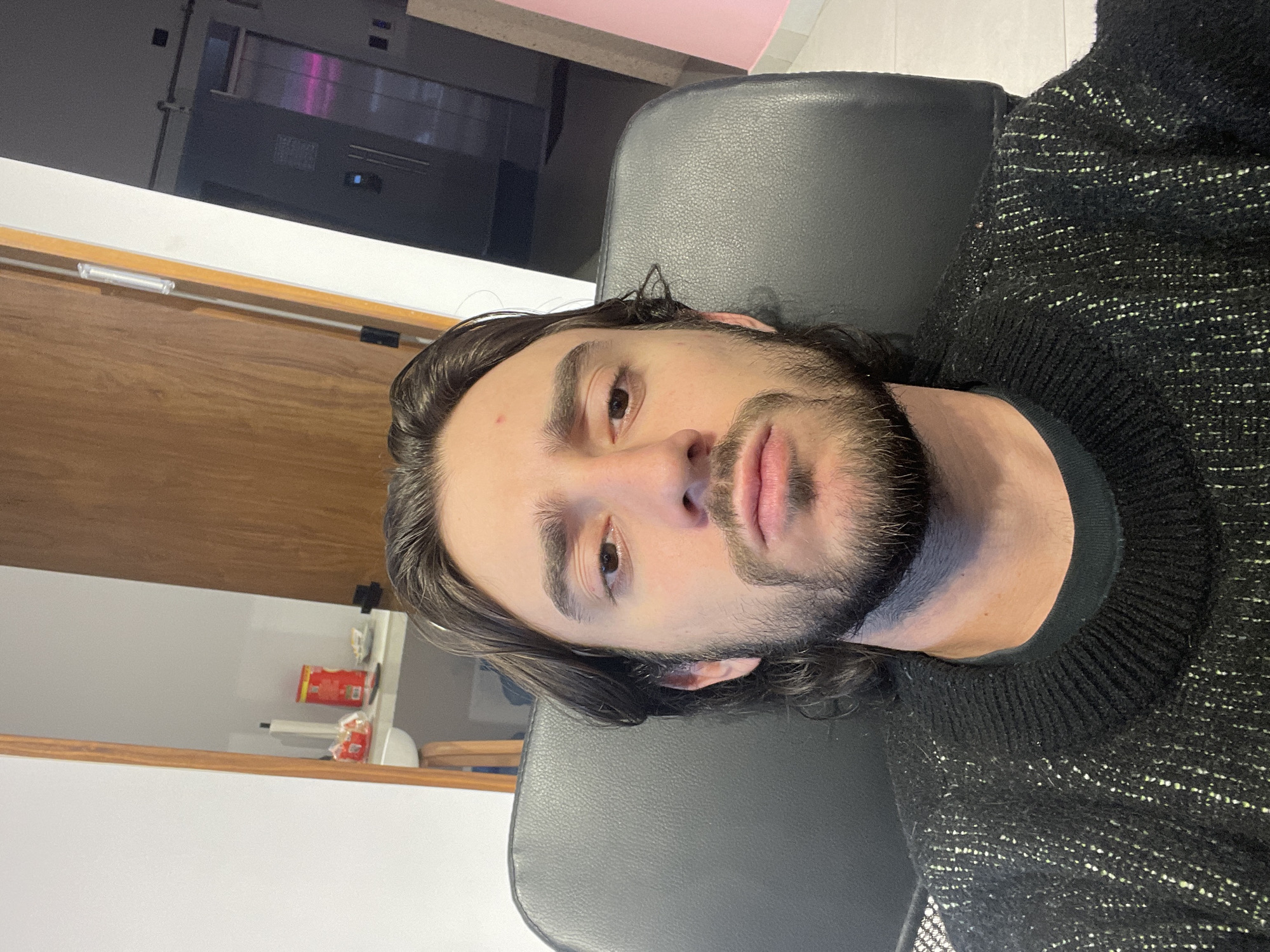 A man with long brown hair, brown eyes, and a beard is looking directly at the camera. He is wearing a black sweater and sitting in a black chair. The background includes a wooden door, a white wall, and a modern appliance. The location appears to be an indoor setting. There is no information about the location in the city of Mexico City, Mexico.