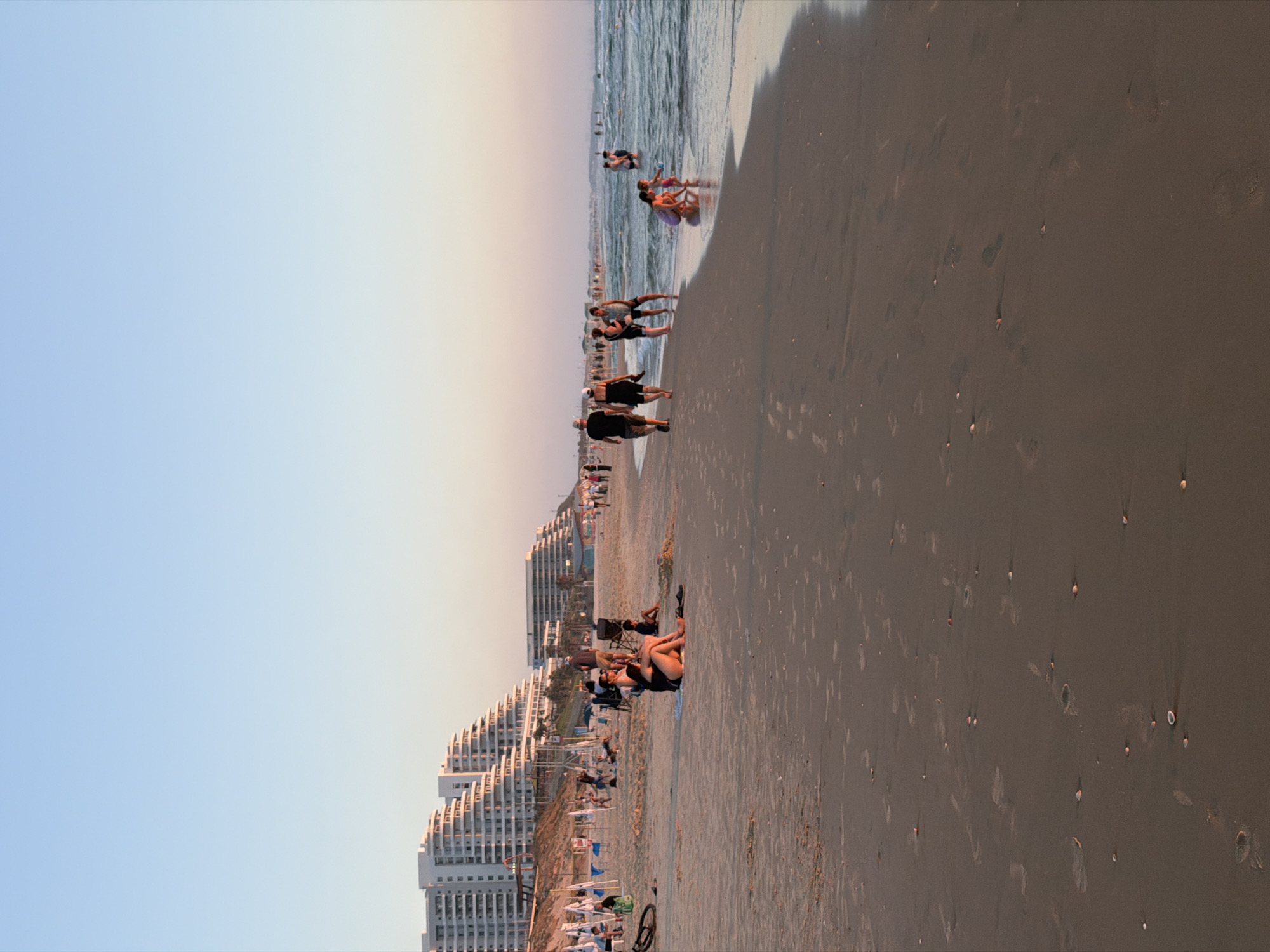 The image depicts a sunset scene at Bat Yam beach in Israel. The sky is a gradient of pale blue to pink. In the background, several modern, multi-story white buildings with stepped facades line the coastline. The beach itself is sandy with numerous footprints and scattered seashells.
In the foreground, people are engaged in various beach activities. To the left, a bicycle is lying on its side. A group of people are sitting and relaxing on the sand. Further down the beach, towards the water, several individuals are walking along the shoreline. A family, including two adults and a child, are interacting with the shallow waves. Others are wading or swimming in the sea. The overall atmosphere appears to be one of leisure and relaxation as the day concludes.