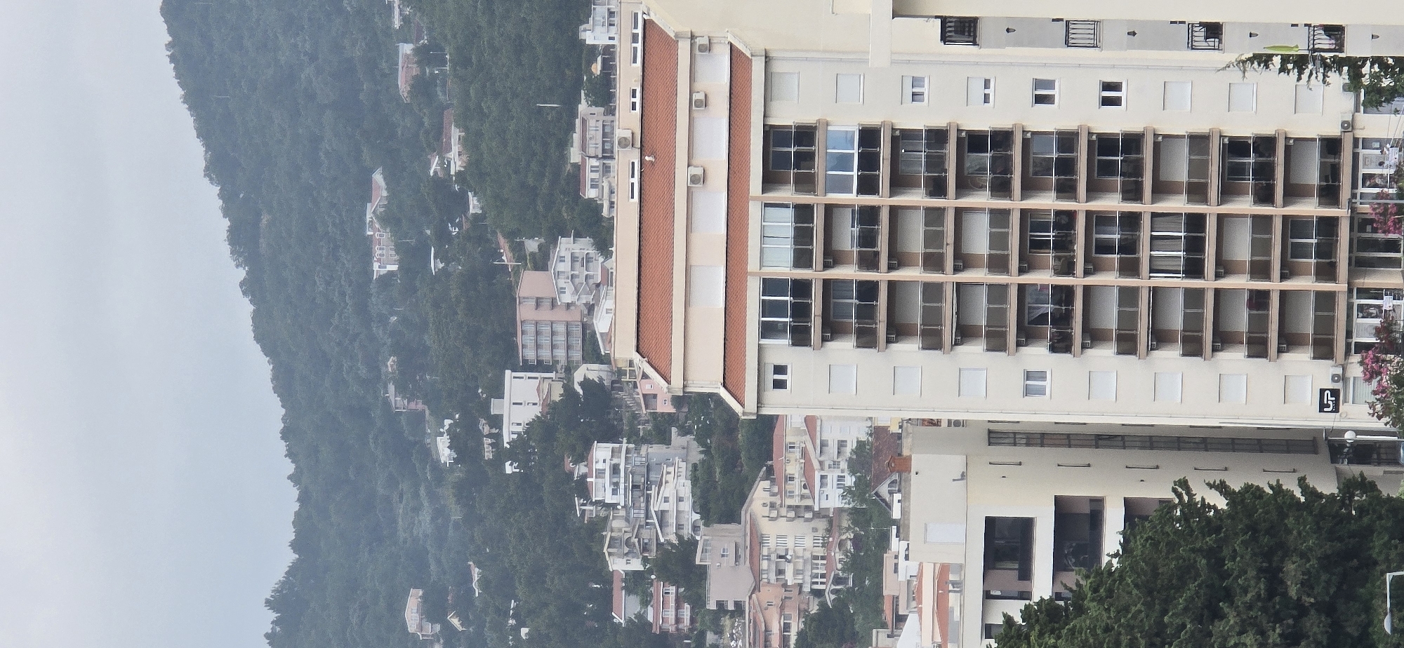 An eye-level shot captures a panoramic view of a multi-story building and a densely forested mountain under an overcast sky, likely in Bar, Montenegro. The prominent feature is a large, light-colored (beige/cream) building occupying the right side of the frame, extending from mid-ground into the foreground. This building features multiple floors with rows of rectangular windows and balconies, many with vertical dividing panels. Sections of its roof are covered with terracotta tiles, and several air conditioning units are visible on ledges and roof areas.
To the left and in the background, a steep, green mountain or hill rises, thickly covered with trees. Numerous smaller residential houses, mostly light-colored, are dotted across the forested slopes, blending into the natural landscape. The sky is a uniform light grey, suggesting an overcast or hazy day, providing soft, diffused lighting across the scene. In the lower right corner of the main building, a black, stylized logo or symbol, possibly resembling an 'S' or a 'G' with an upward-pointing arrow or loop, is visible on the facade. No human activity or specific events are discernible.