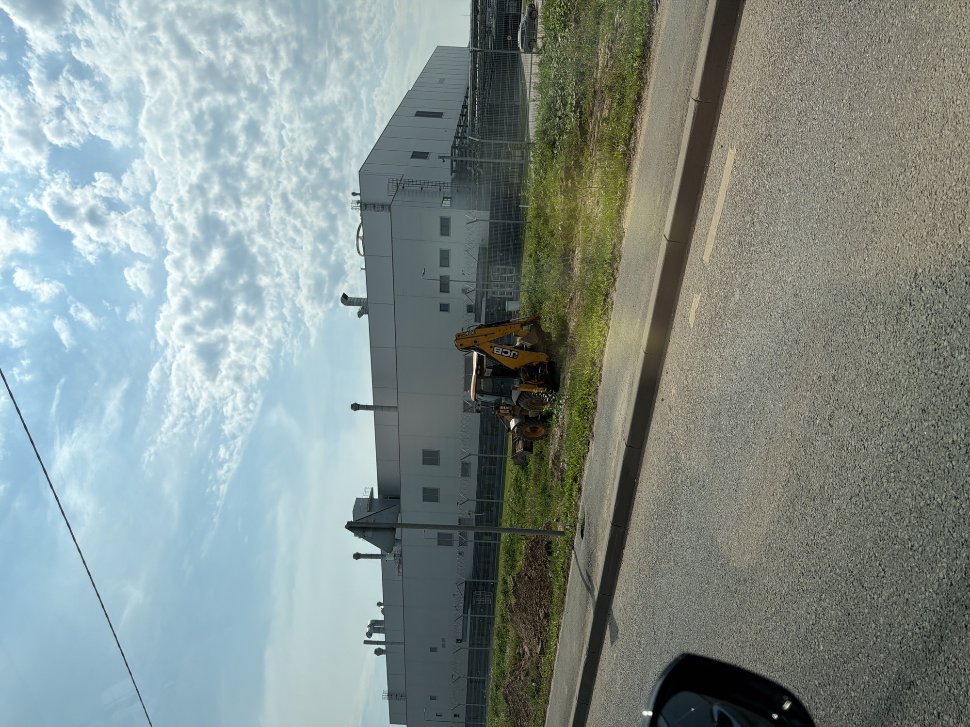 A yellow JCB backhoe loader is parked on a patch of grass next to a pile of dirt, in front of a large, modern industrial building with a white facade and several ventilation pipes on the roof. A barbed wire fence runs along the perimeter of the building. A road with a curb runs in the foreground, and a portion of a car's side mirror is visible in the bottom left corner. The sky is blue with scattered white clouds. The scene appears to be in an industrial area, likely on the outskirts of a city. The location is identified as Калининец-2 Снт, Russia.