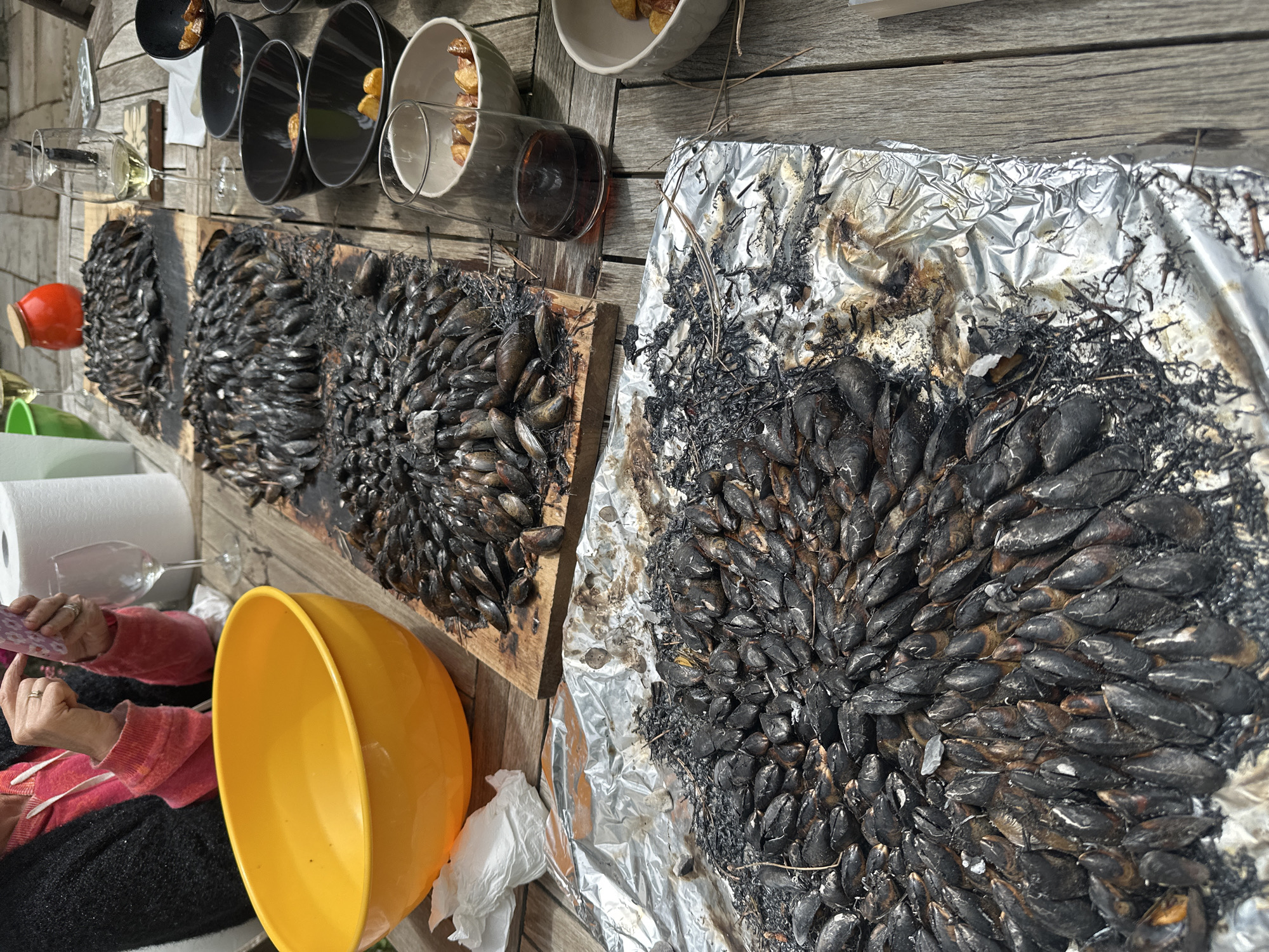 The image captures an outdoor food preparation scene in La Couarde-sur-Mer, France. Several wooden planks form a table surface on which the main subject of the image rests. Three wooden planks form a tabletop. Two sets of cooked mussels dominate the view. The first set is arranged on a wooden board, the second on a piece of aluminum foil. Next to the mussels, six ceramic bowls with fried potatoes are visible, along with three empty wine glasses. In the lower-left corner, a yellow plastic bowl and a partially visible person in a red sweater and black pants suggest active preparation. The background consists of a light-colored wooden wall.