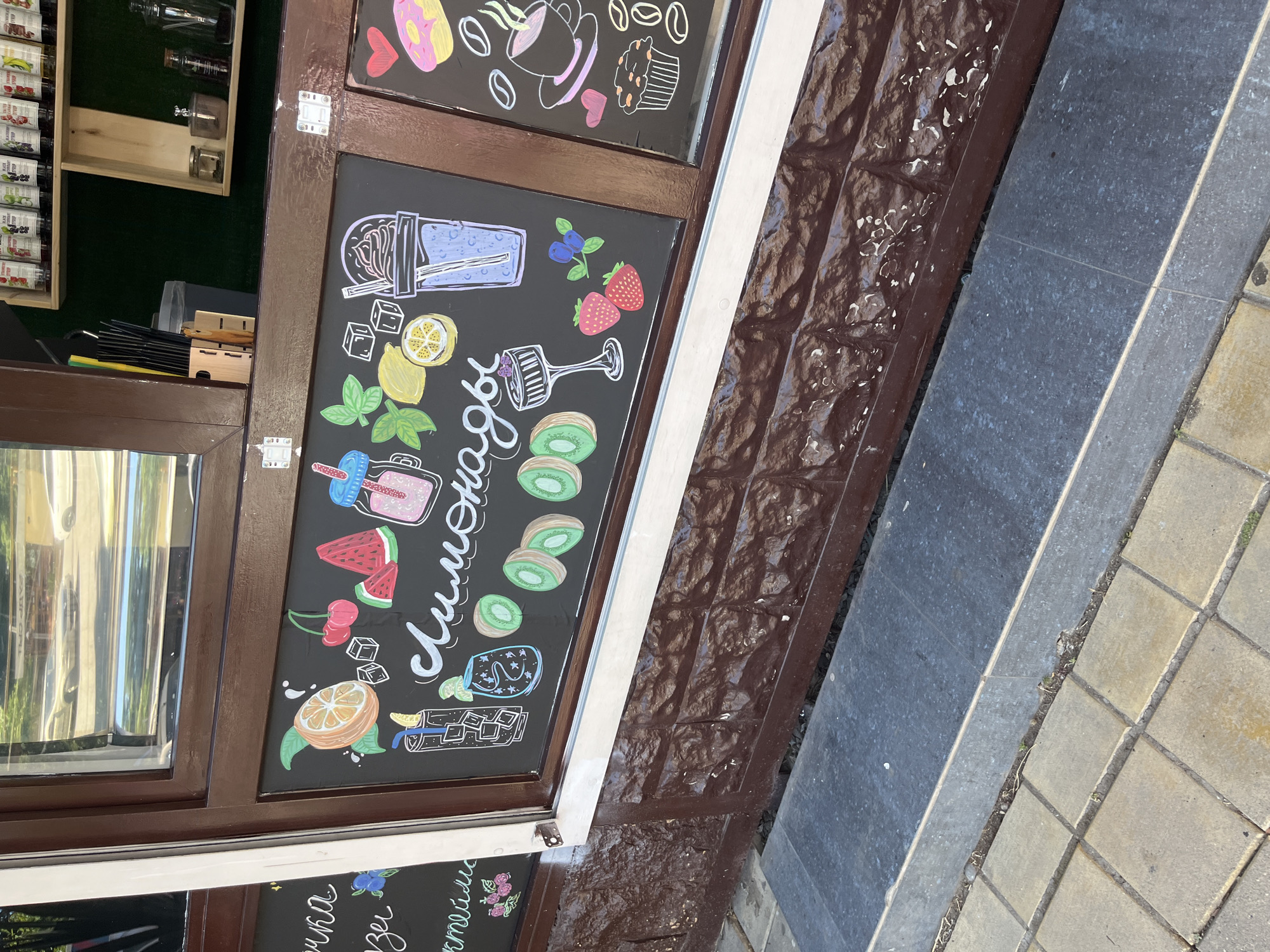 The image shows a close-up of a sidewalk window display with a chalkboard menu. The chalkboard is decorated with colorful chalk drawings of fruits, drinks, and pastries. Specifically, there are drawings of an orange, cherries, watermelon slices, kiwi slices, strawberries, blueberries, ice cubes, a glass of iced tea or lemonade, a smoothie, a dessert in a glass, and a large cup with a straw. The word "Лимонады" (Lemonades) is written in cursive script across the chalkboard. To the right, on another section of the chalkboard, are drawings of a coffee cup with steam, a cupcake, coffee beans, and a heart shape. The window frame is dark brown. The window itself reflects a street scene with cars and trees. Below the window is a brown, textured wall and a dark gray step leading to yellow square pavers. The overall scene suggests a cafe or a beverage stand in an outdoor setting. The mention of Sochi, Russia in the prompt is not explicitly evident from the visual information alone.