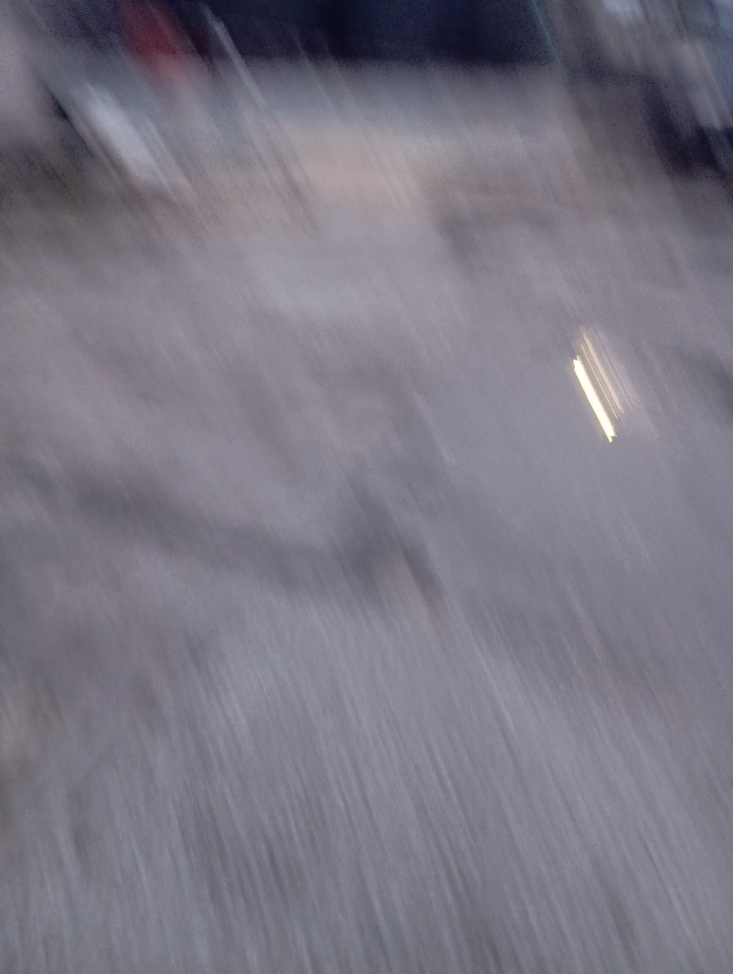 The image is a blurred, low-light photograph with a diagonal motion blur. It appears to be an outdoor scene, possibly a street or an open area. There are faint suggestions of structures or vehicles in the upper portion of the image, with some indistinct colors that could be red or dark blue. A bright, elongated light source is visible in the mid-right section, appearing as streaks due to the motion blur. The ground or surface is a muted, greyish tone with a texture suggestive of gravel or rough pavement. The overall impression is one of movement or a hasty capture in dim lighting conditions. Without more distinct features, pinpointing a specific location in Port Harcourt, Nigeria is not possible.