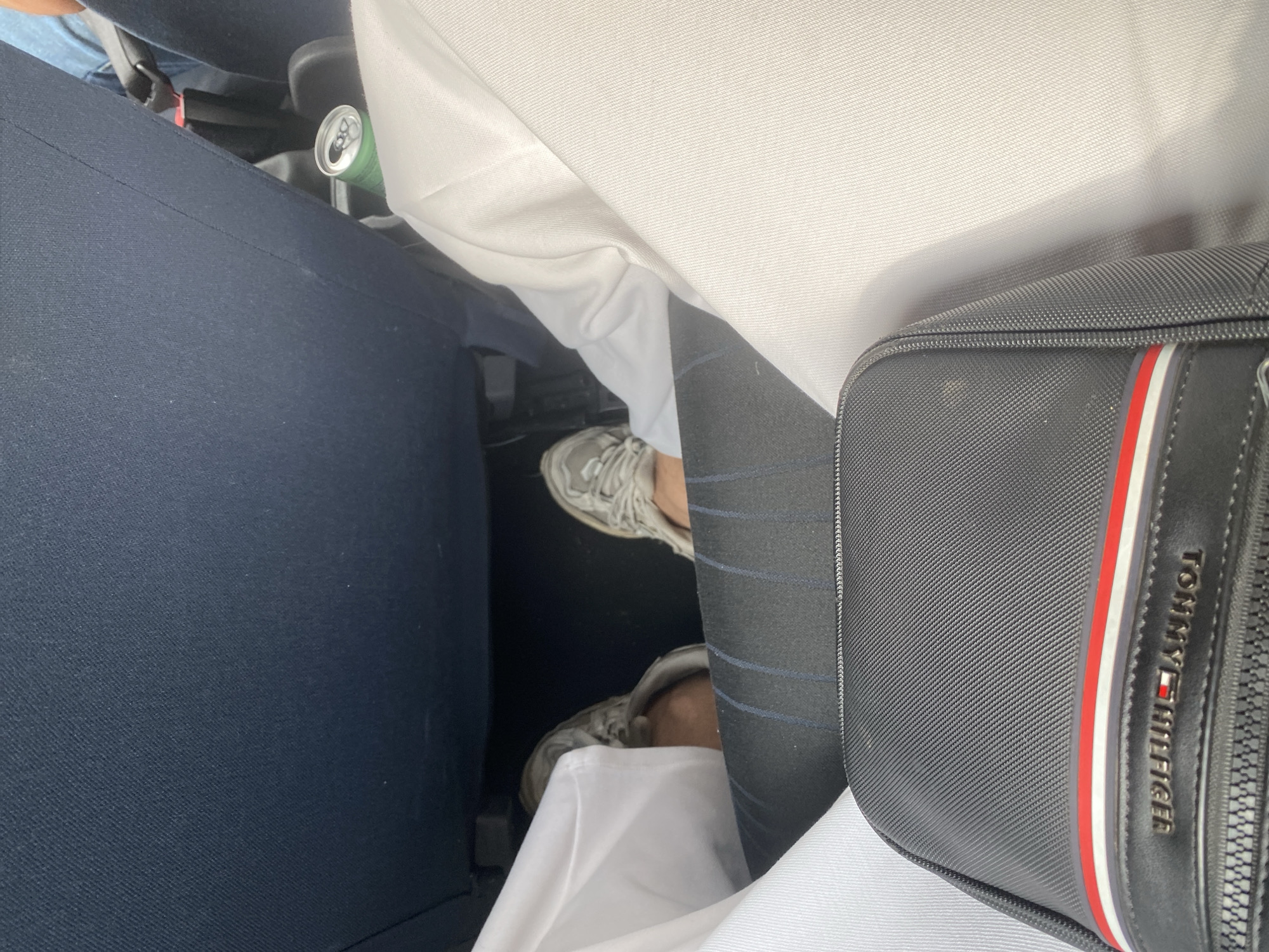 The image shows a low-angle view of the legs and lower torso of a person sitting in what appears to be a car or airplane. The person is wearing grey athletic shoes and white clothing, possibly a thawb or robe. A black Tommy Hilfiger bag is in the foreground, with its logo visible. The seating appears to be dark blue fabric. A partially opened can of a beverage is resting in a cupholder or on the floor to the right. The scene is indoors and the lighting is neutral. There is no visible indication of the city of Weyhe, Germany.