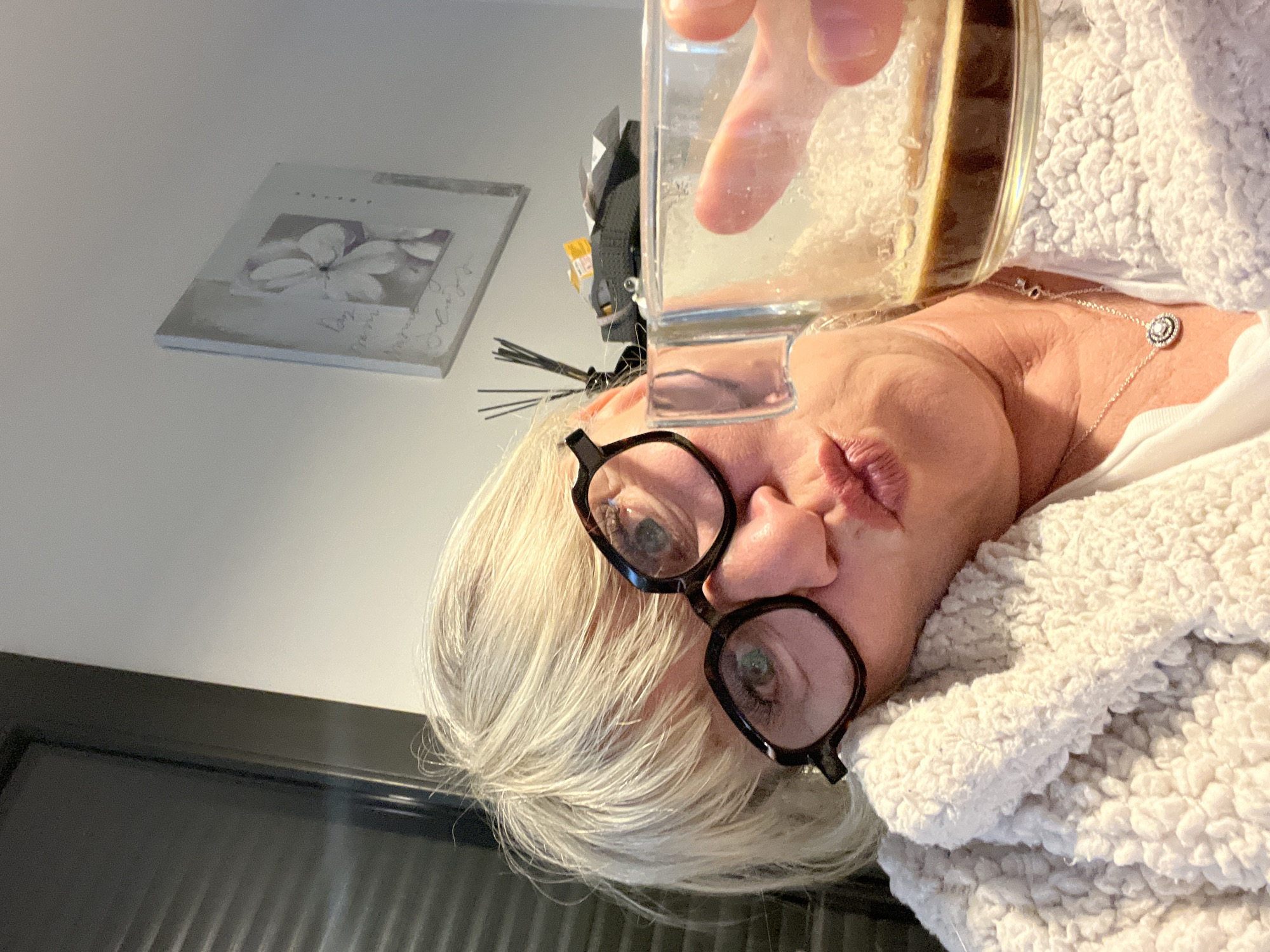 A woman with short, grey hair and black framed glasses is holding a glass of liquid close to the camera. She is wearing a white top and a light-colored, fluffy jacket. Her lips are pursed. A silver necklace is visible around her neck. Behind her is a white wall with a framed picture of a flower on it. To the left is a dark colored door. There is also a black wall decoration, a clock, and aroma diffusers behind the woman. The image does not contain enough information to confirm if the location is Chambœuf, France.