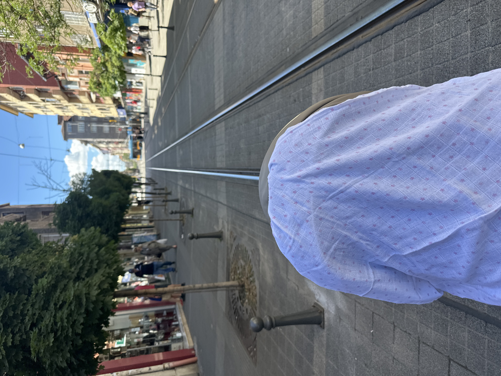 This is a POV shot from a stroller, looking forward down a city street with tram tracks. The stroller is covered with a light blue blanket with a subtle pink star pattern. To the left of the tram tracks, there's a sidewalk lined with trees, shops, and people walking. To the right of the tracks, the street continues with more buildings and pedestrians. In the distance, a tram is visible. The sky is blue with some clouds, and the sun is shining, casting shadows. The overall scene depicts a typical urban environment on a bright day.