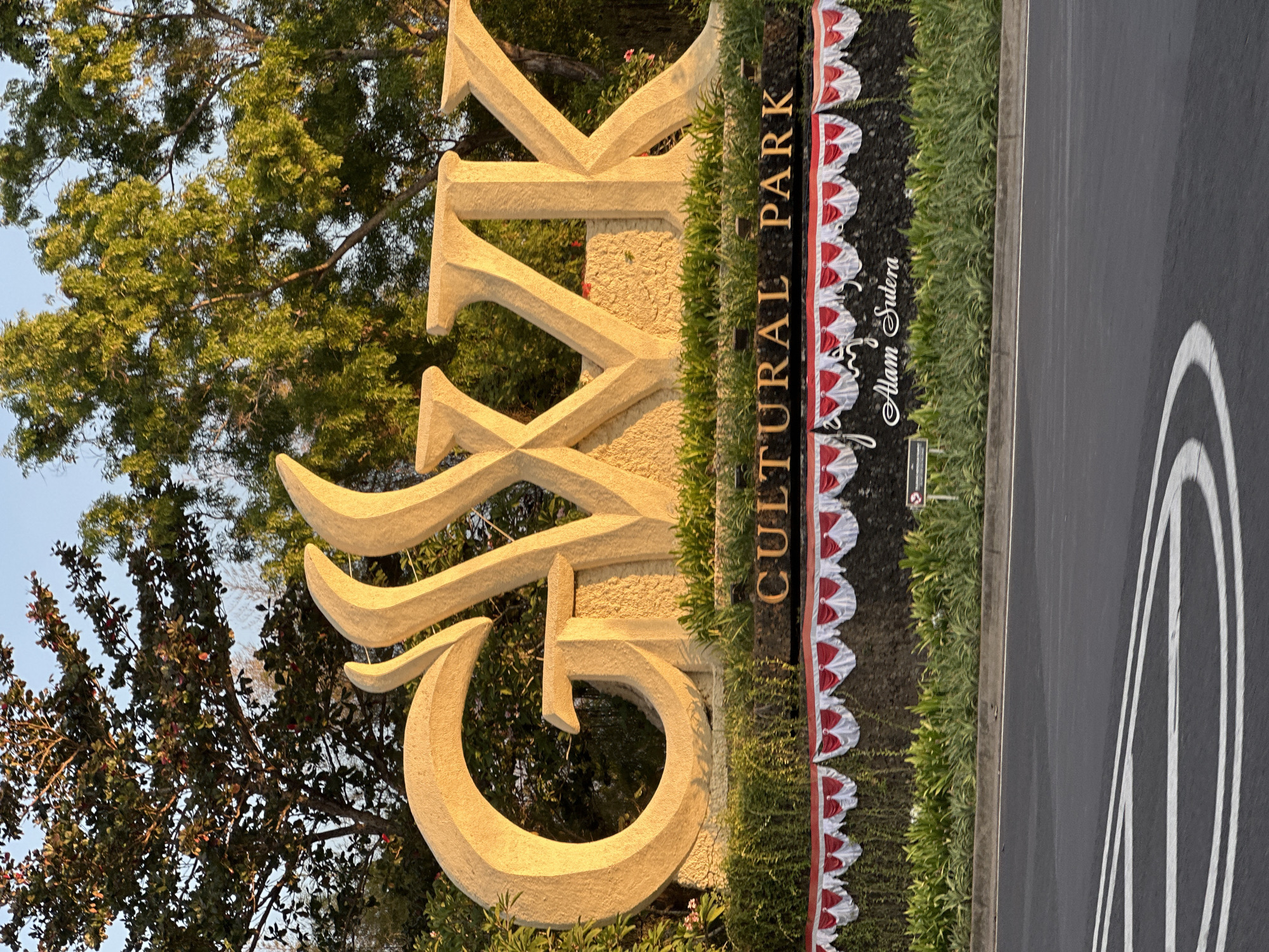 The image shows the large, stone letters "GWK" at the entrance to the Garuda Wisnu Kencana Cultural Park in Kuta Selatan, Bali, Indonesia. The letters are illuminated by the warm, golden light of the setting sun. Below the letters, a dark stone plaque reads "CULTURAL PARK" in gold lettering, with a decorative border of red and white fabric draped above it. Further down, in a stylized font, the words "Alam Sutera" are visible. The park is surrounded by lush green foliage and trees. In the foreground, a road with white markings is visible. The sky above is a clear, pale blue.