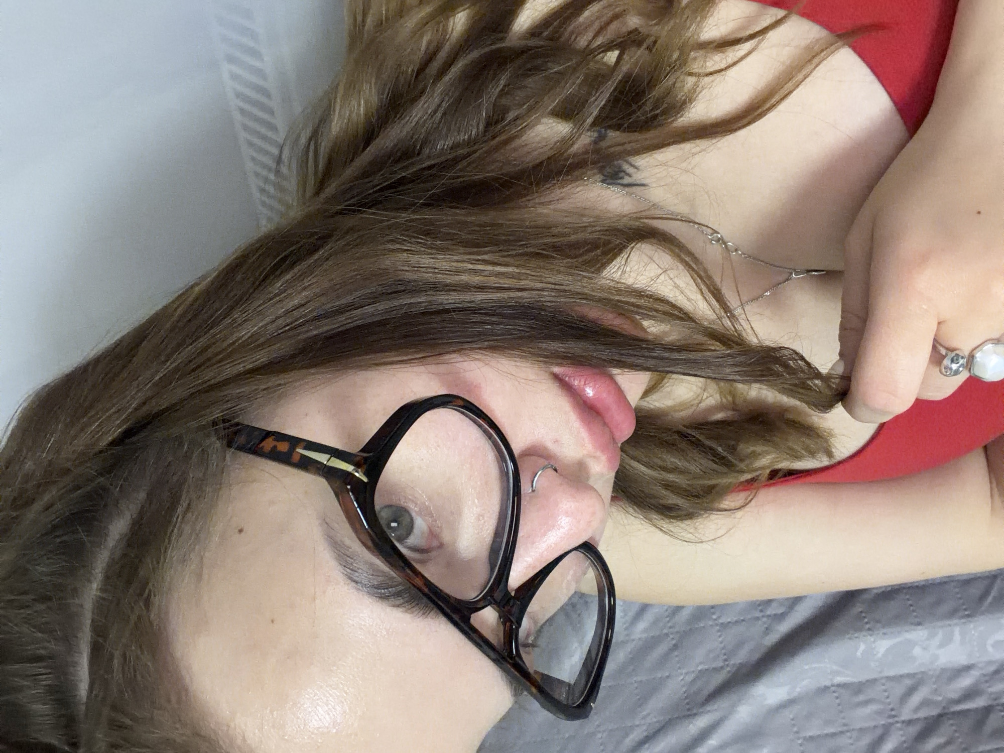 A woman with light skin, brown hair, and gray eyes is lying on a gray quilted surface, possibly a bed. She is wearing eyeglasses with a tortoise shell pattern and a red top. She has a silver nose ring and a silver necklace. A ring is visible on her hand. The location is not evident from the image.