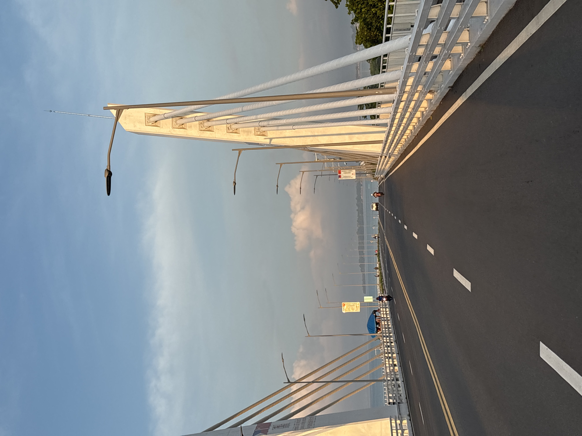 This image captures a view of the Francisco Bangoy Bridge, also known as the Davao-Samal Bridge, in Davao City, Philippines, likely during golden hour as indicated by the warm lighting on the bridge structure. The perspective is from the roadway, looking ahead over the bridge's expanse.
The bridge itself is a prominent cable-stayed structure with large, white pylons supporting the cables that fan out to hold the road deck. The roadway is asphalt, marked with double yellow lines and dashed white lines, indicating multiple lanes. Streetlights are spaced along the bridge's length.
On the left side of the road, there's a white pylon with several large cables descending from it. A blue tent is visible on the pedestrian walkway on the left, with several people gathered underneath it. A person on a motorcycle is visible in the foreground on the left lane, heading away from the camera. Further down the road, more vehicles and people are present.
On the right side of the road, another white pylon mirrors the one on the left. The railing of the bridge, made of metal bars, runs along the side of the roadway. The background shows a body of water, likely the Davao Gulf, with a distant shoreline and lush green trees. The sky is a clear blue with some scattered, soft clouds, tinged with the warm colors of the setting or rising sun. The overall scene depicts a functional piece of infrastructure within a scenic coastal environment.