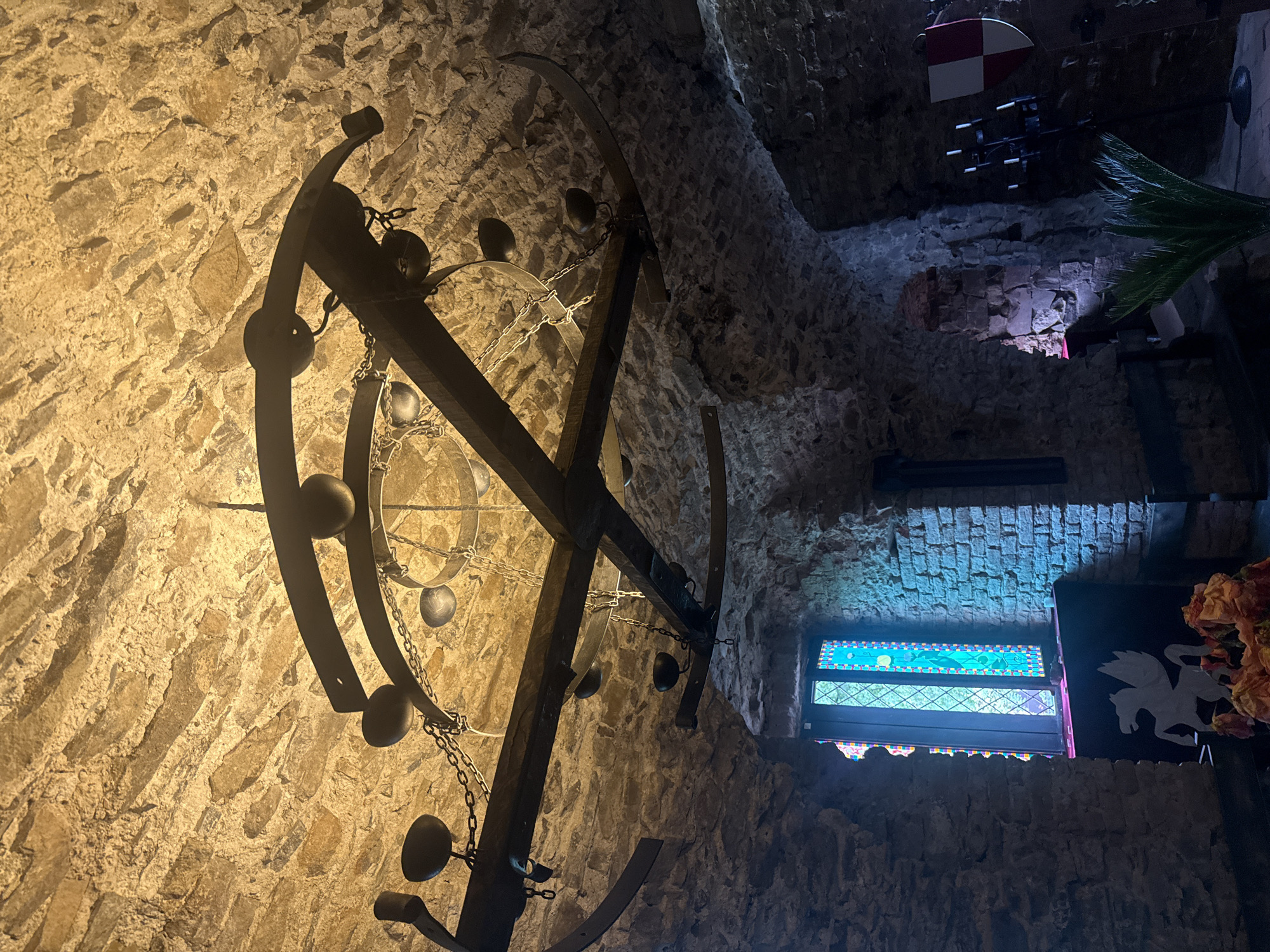 The image captures the interior of St. Miklós Castle in Chynadiiovo, Ukraine, showcasing a room with rough stone walls and a unique, ornate chandelier. The chandelier is constructed of metal, featuring a wheel-like structure with decorative metal balls and chains hanging from it. Light shines upon it.
In the background, a section of the castle's interior is visible, including what seems to be another lighting fixture or decorative item hanging near the ceiling. A stained glass window is visible.