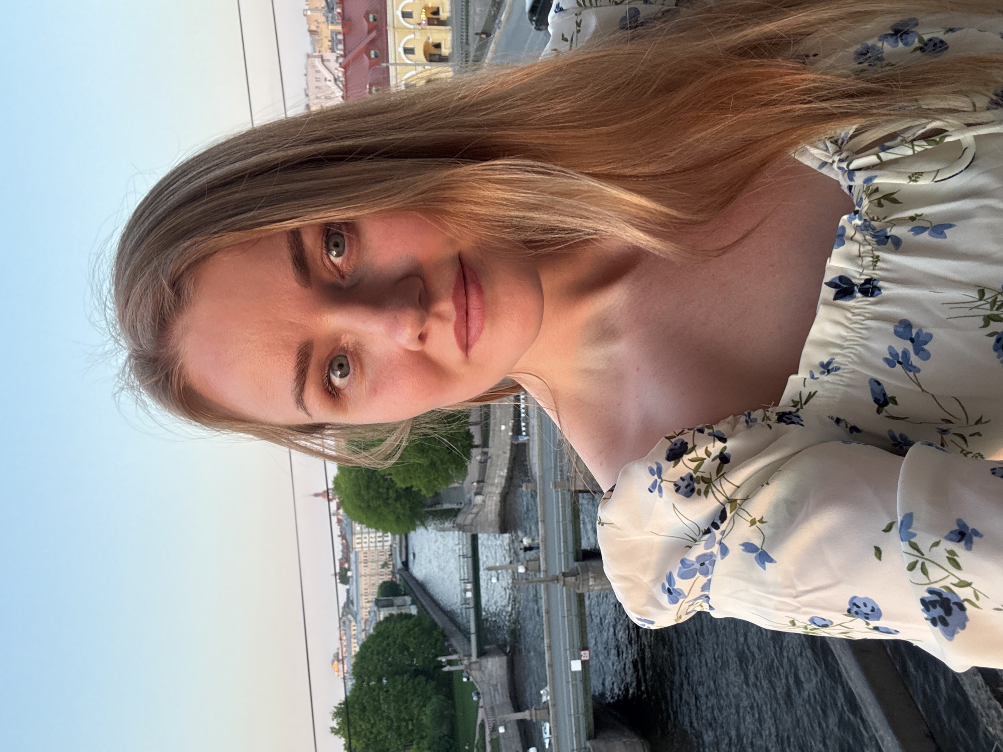 A young woman with light skin and long blonde hair poses for a close-up selfie. She wears a white floral-patterned top. The background shows a section of Saint Petersburg, Russia, featuring a bridge over the Neva River and buildings along the riverbank. Trees are visible along the embankment. The sky is a pale blue, suggesting either dawn or dusk.