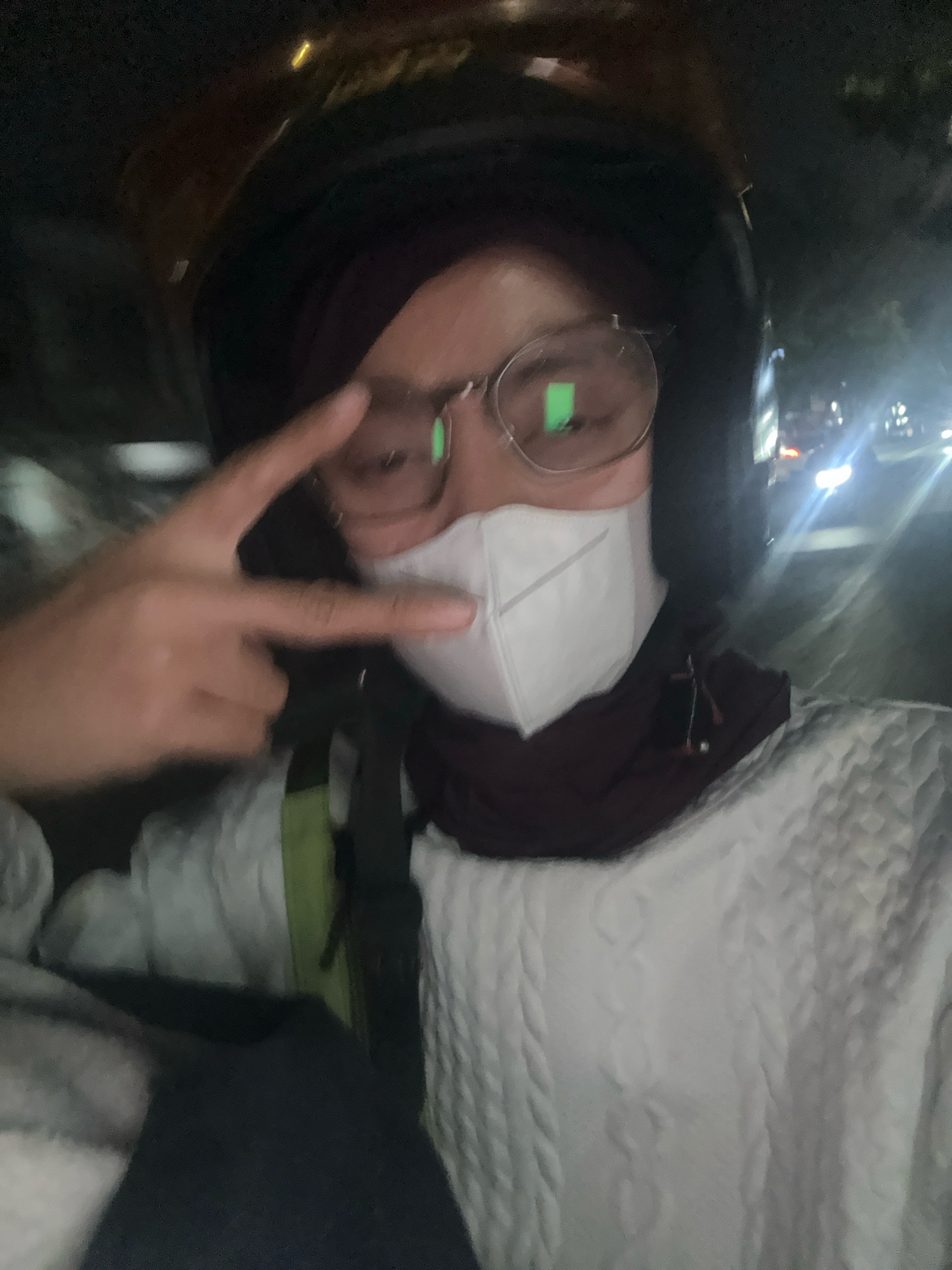 A selfie taken at night shows a person wearing a helmet, glasses, and a white face mask. The person is making a peace sign with their right hand. They are wearing a dark headscarf and a white, textured sweater. The background is dark with blurred lights, suggesting an outdoor or vehicle setting, possibly in a city. The green tint on the glasses suggests a light source reflecting off them. The image is low-resolution and slightly blurry, consistent with a nighttime smartphone photo. The context of Bandung City, Indonesia, is not explicitly visible in the image itself, but is provided as external information.