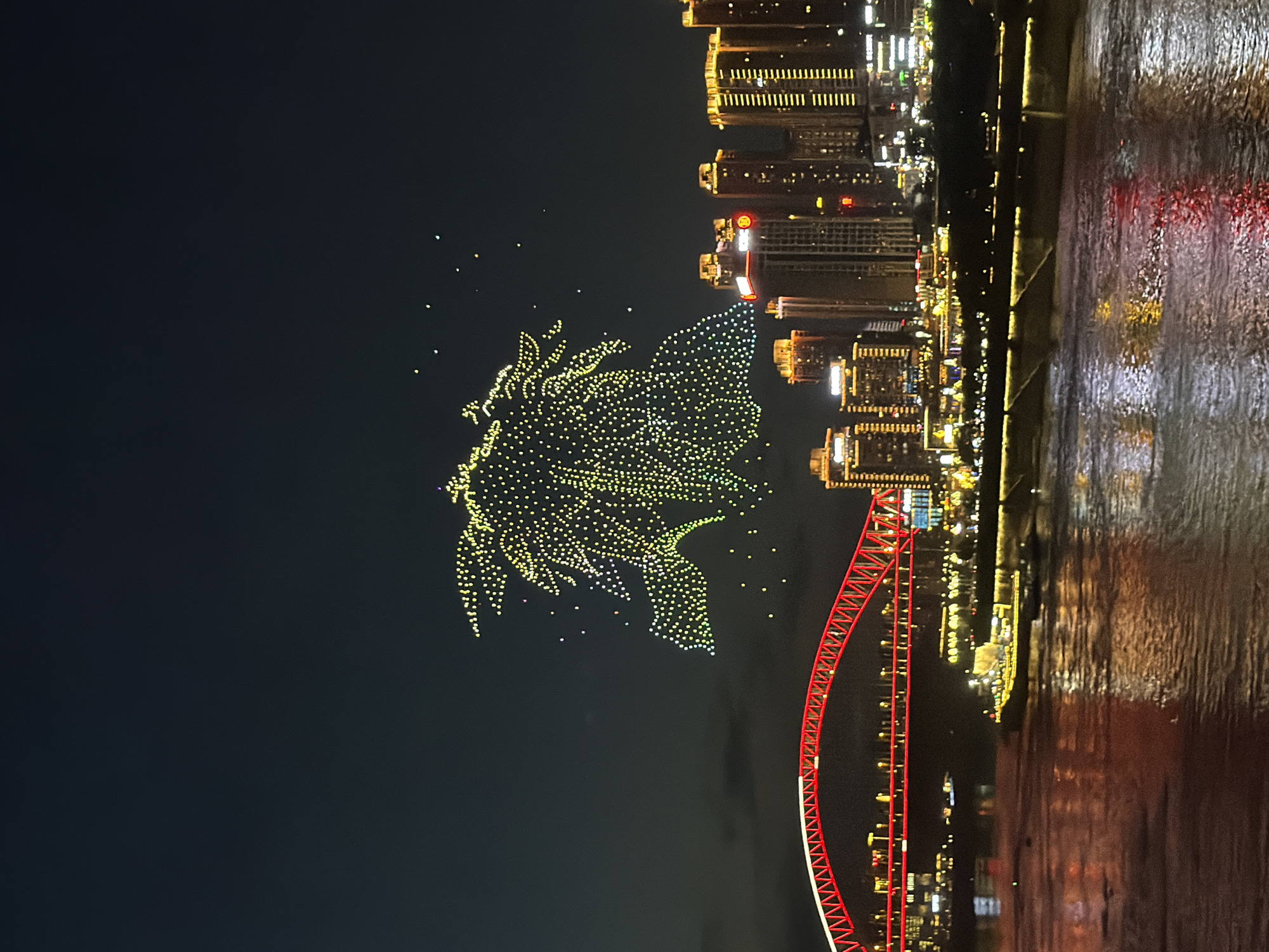 The image captures a nighttime cityscape, likely in the Yuzhong District of China, illuminated by a drone light show and city lights. In the sky, a large, intricate formation of yellow and green lights, resembling a stylized peacock or mythical creature, dominates the upper portion of the frame. Below the drone display, a vibrant red bridge with a curved arch is prominently featured, spanning across a body of water. The bridge is illuminated with red lights along its structure. On the far bank of the water, a cityscape of tall buildings, many of which are lit with yellow and orange lights, forms the background. A large barge or ship, also lit, is visible on the water in front of the city skyline. The water itself reflects the lights from the bridge and the city, creating shimmering patterns. The overall atmosphere is one of a festive and technologically advanced urban environment at night.